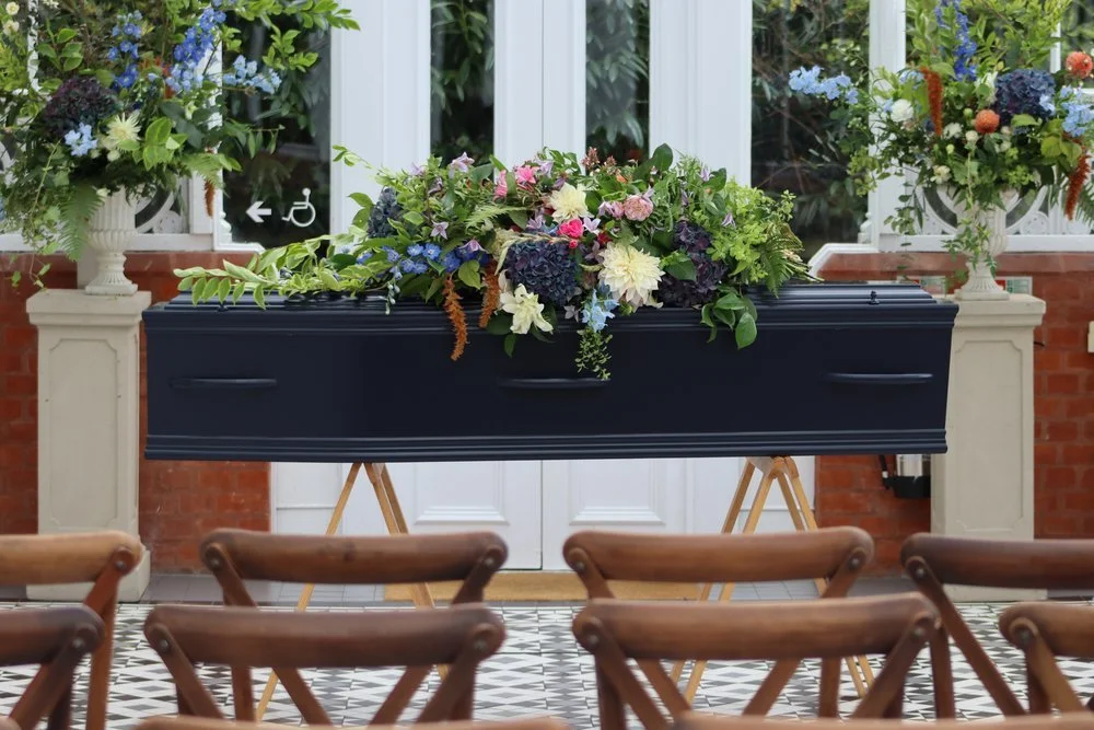 A flower-covered coffin inside the Horniman Conservatory with rows of chairs prepared for a funeral