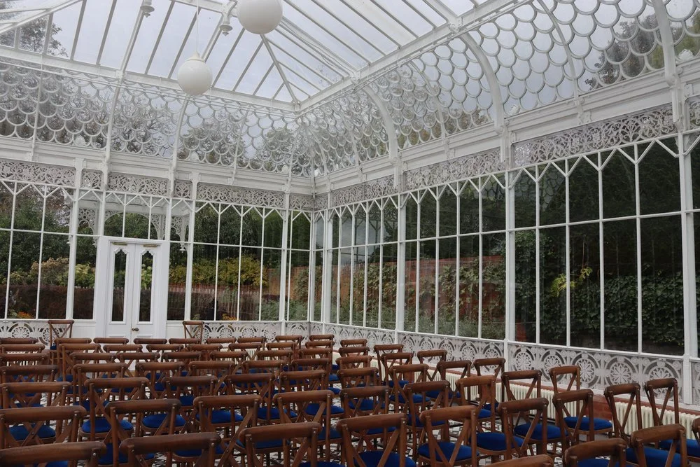 Rows of wooden ceremony chairs beneath the intricate glass roof of the Horniman Conservatory