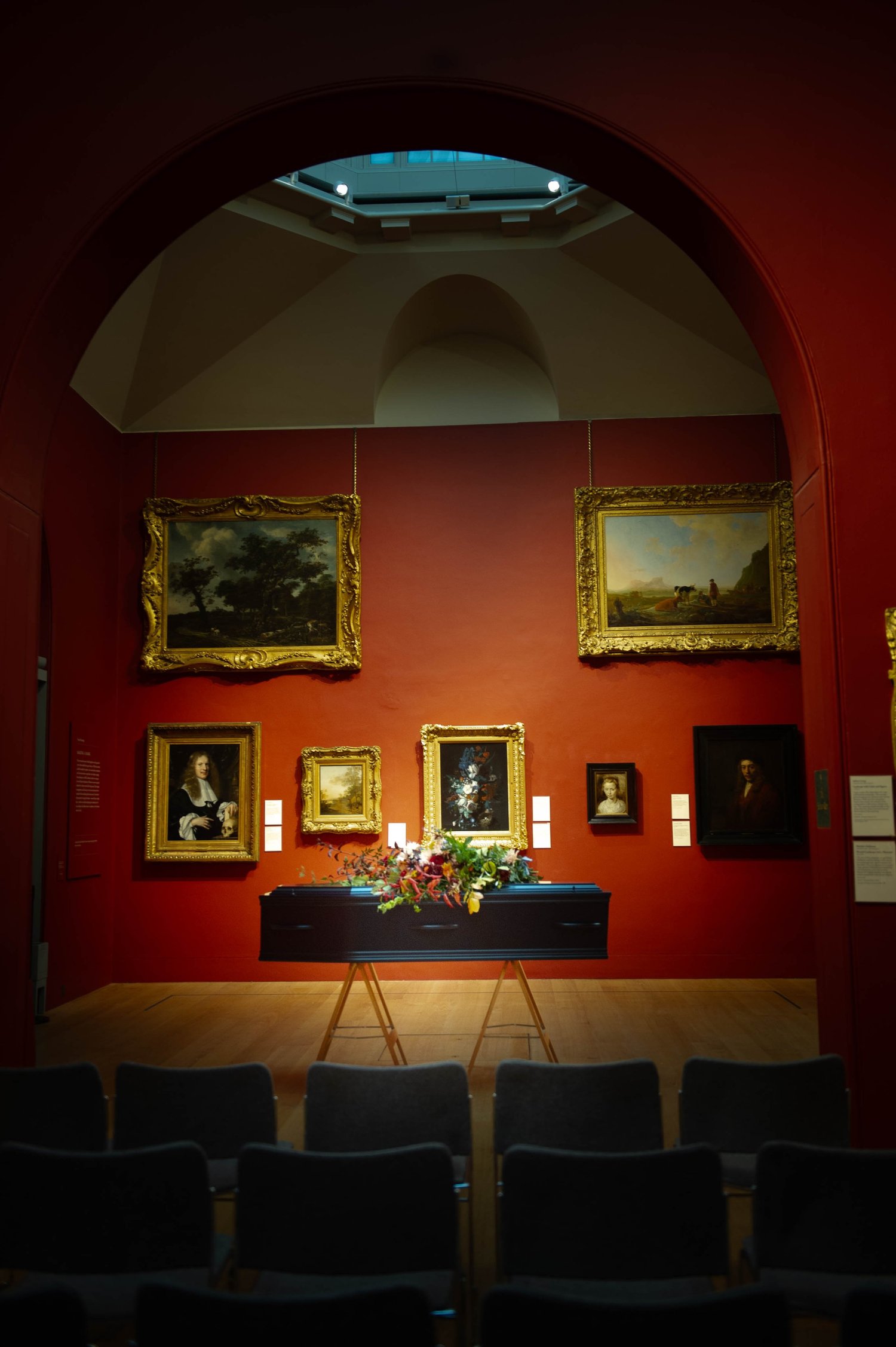 A flower-covered coffin beneath historic paintings in a red-walled gallery at Dulwich Picture Gallery