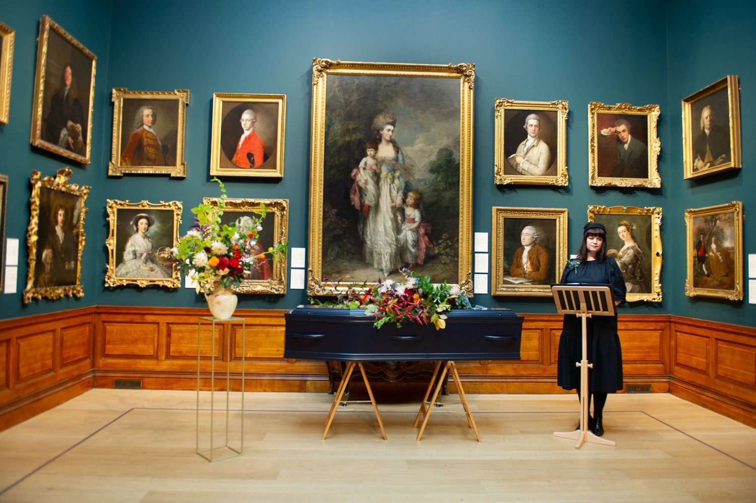 A speaker at a lectern beside a flower-covered coffin in Dulwich Picture Gallery