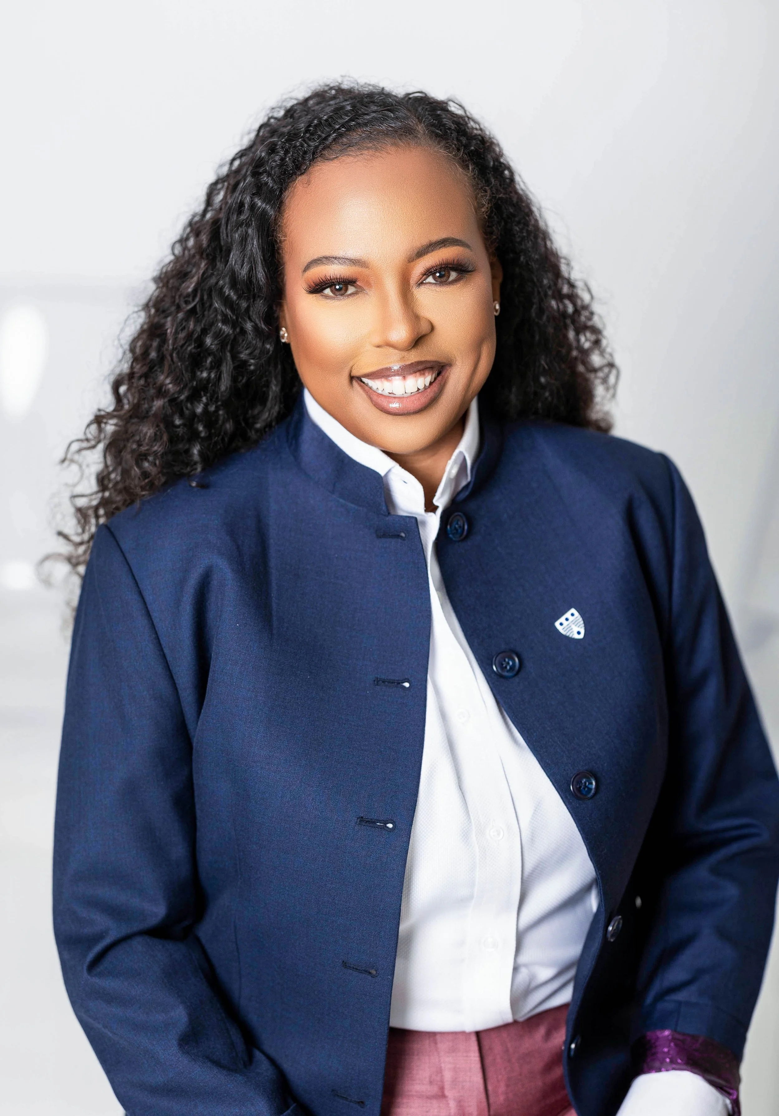 Professional woman with curly black hair, wearing a navy blazer, white shirt, and pink pants, smiling at the camera.