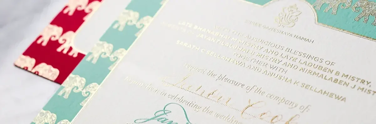 Close up of Hindu wedding invite with gold foil elephant pattern border with a teal and bright pink background and elegant gold typography