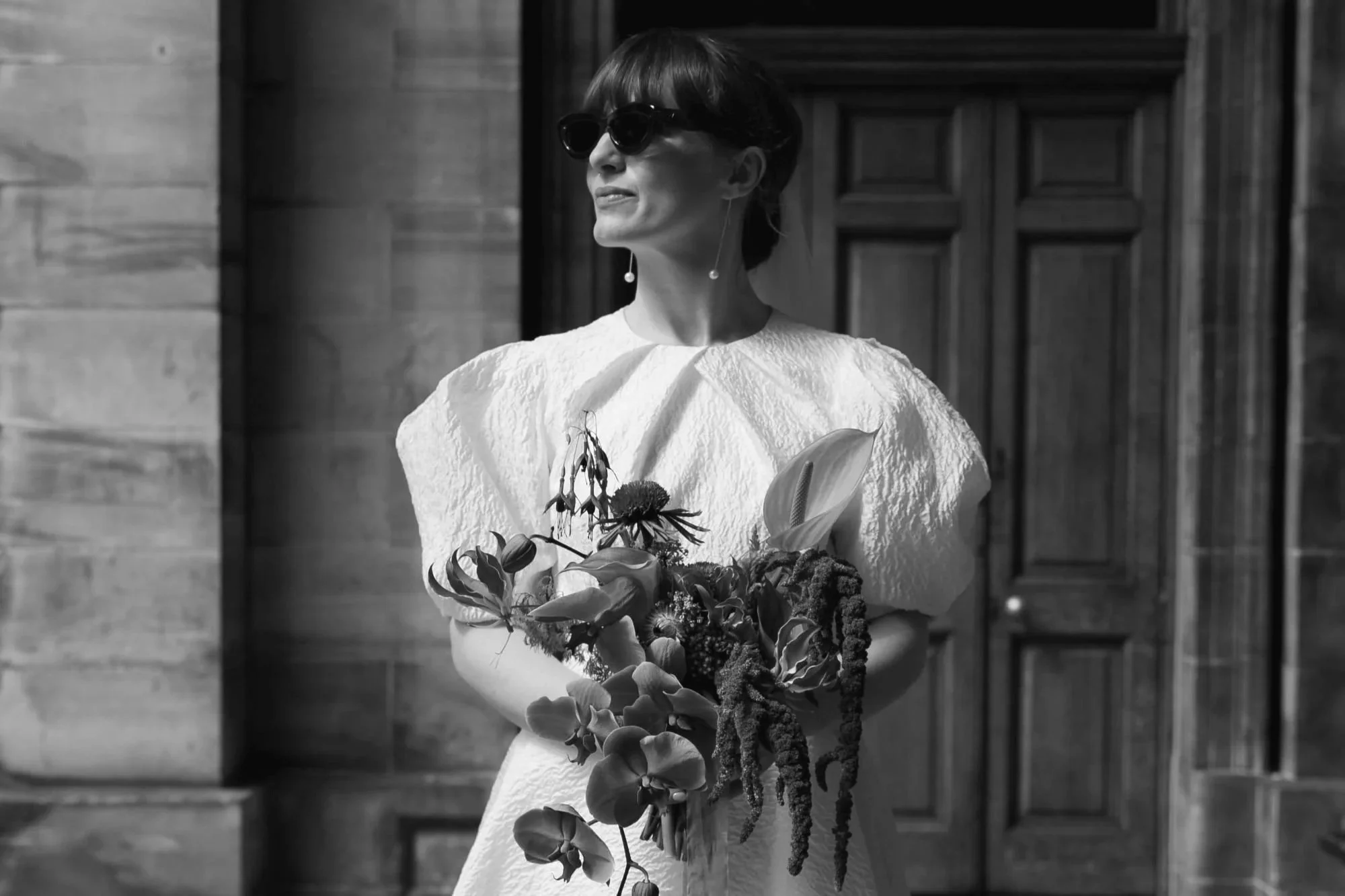 A woman wearing sunglasses and earrings, holding a bouquet of flowers, standing outside a building with wooden doors.