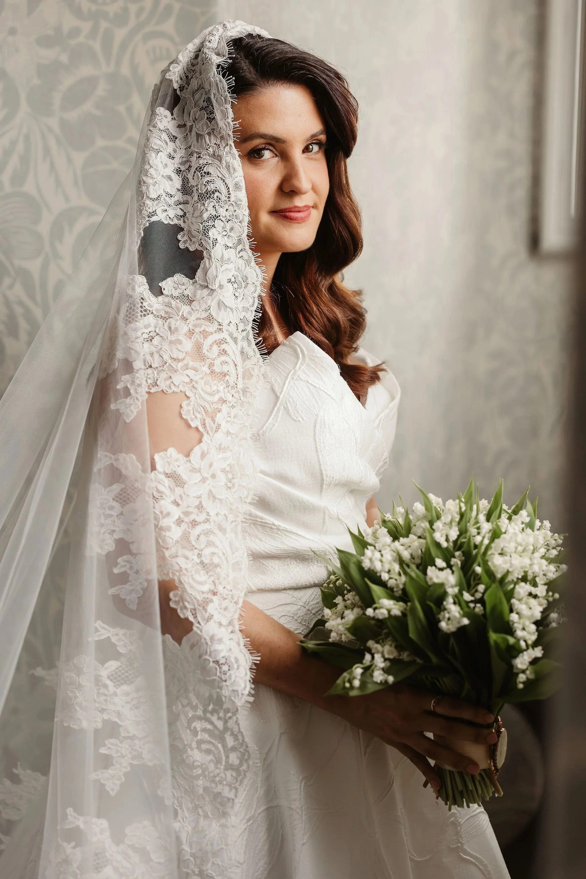 A bride with dark brown wavy hair holding a bouquet of white flowers with green leaves, wearing a white dress and a lace veil, standing near a window with patterned wallpaper in the background.