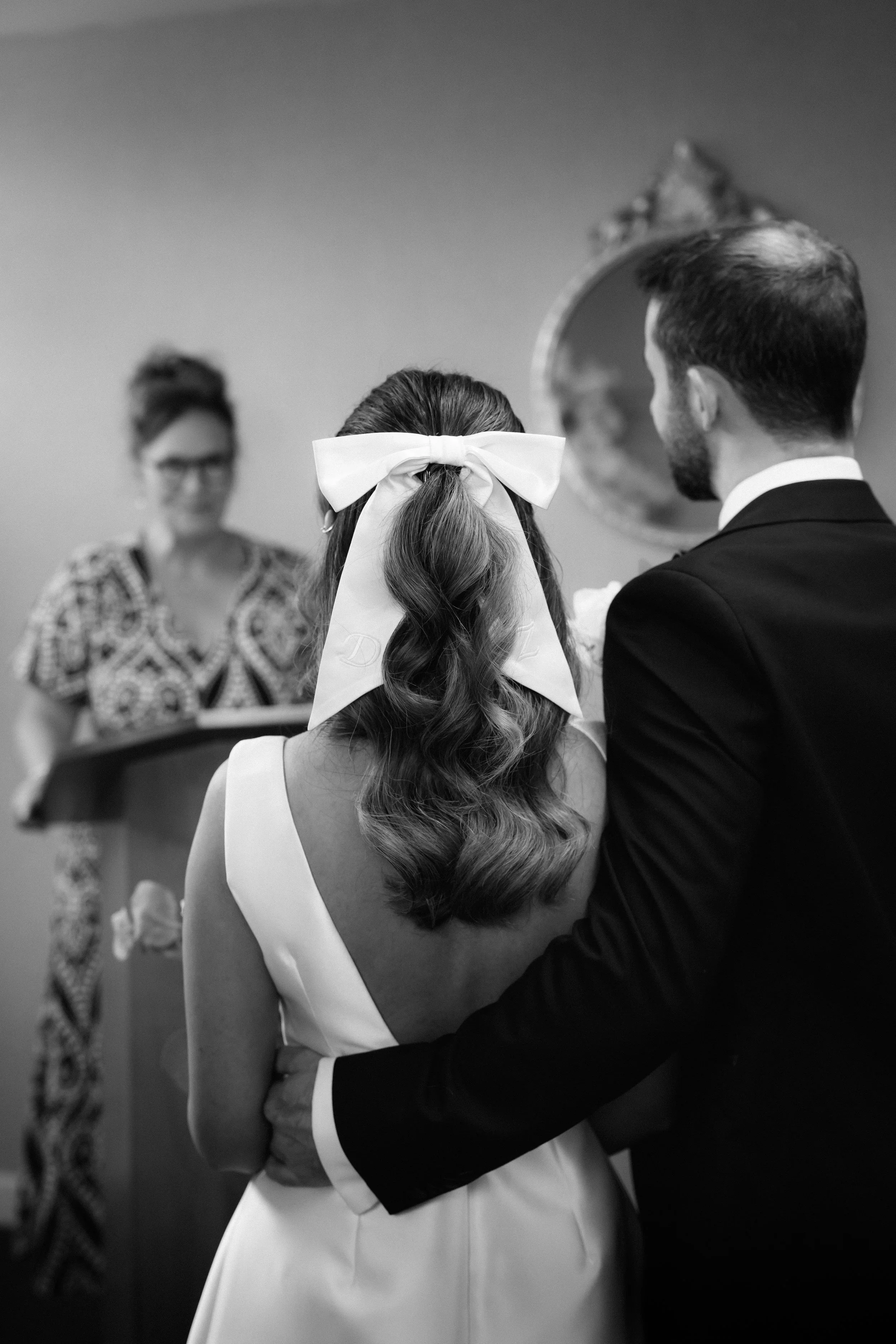 A wedding ceremony with a bride and groom standing together, the bride has a large bow in her hair, and an officiant or speaker in the background.