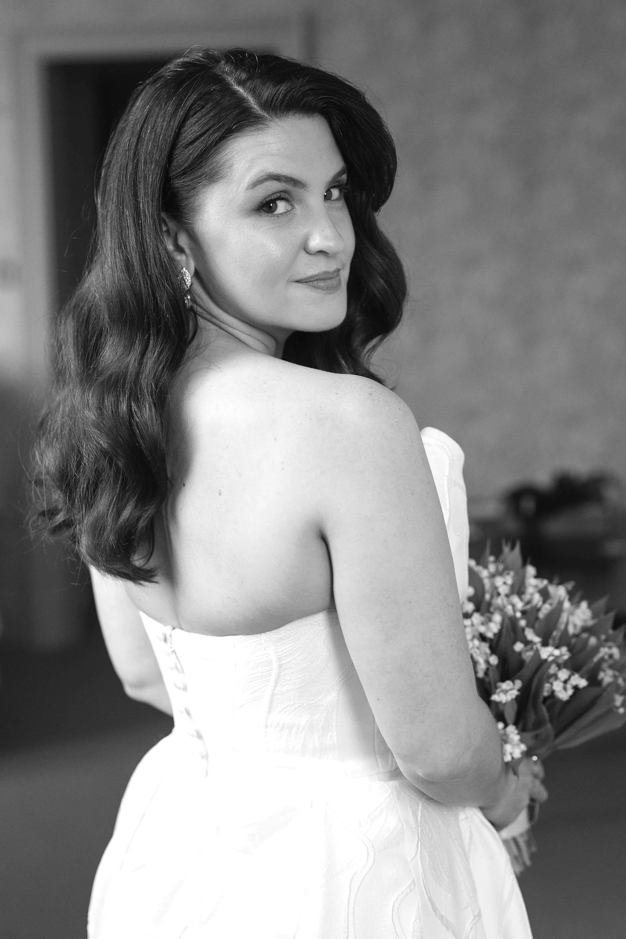 A woman with long, wavy hair wearing a strapless dress, holding a bouquet of flowers, and looking over her shoulder.