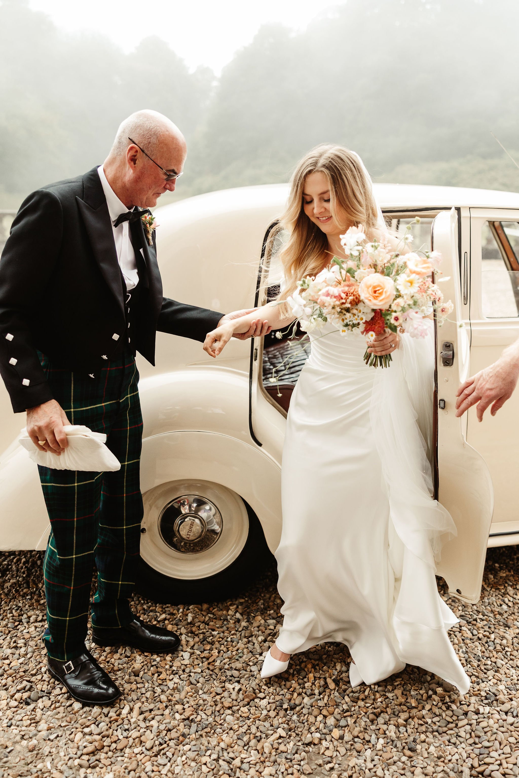 A bride in a white wedding dress holding a bouquet is stepping out of a vintage car, assisted by an older man dressed in a tuxedo with tartan kilt. The scene is set outdoors on a gravel surface with foggy greenery in the background.