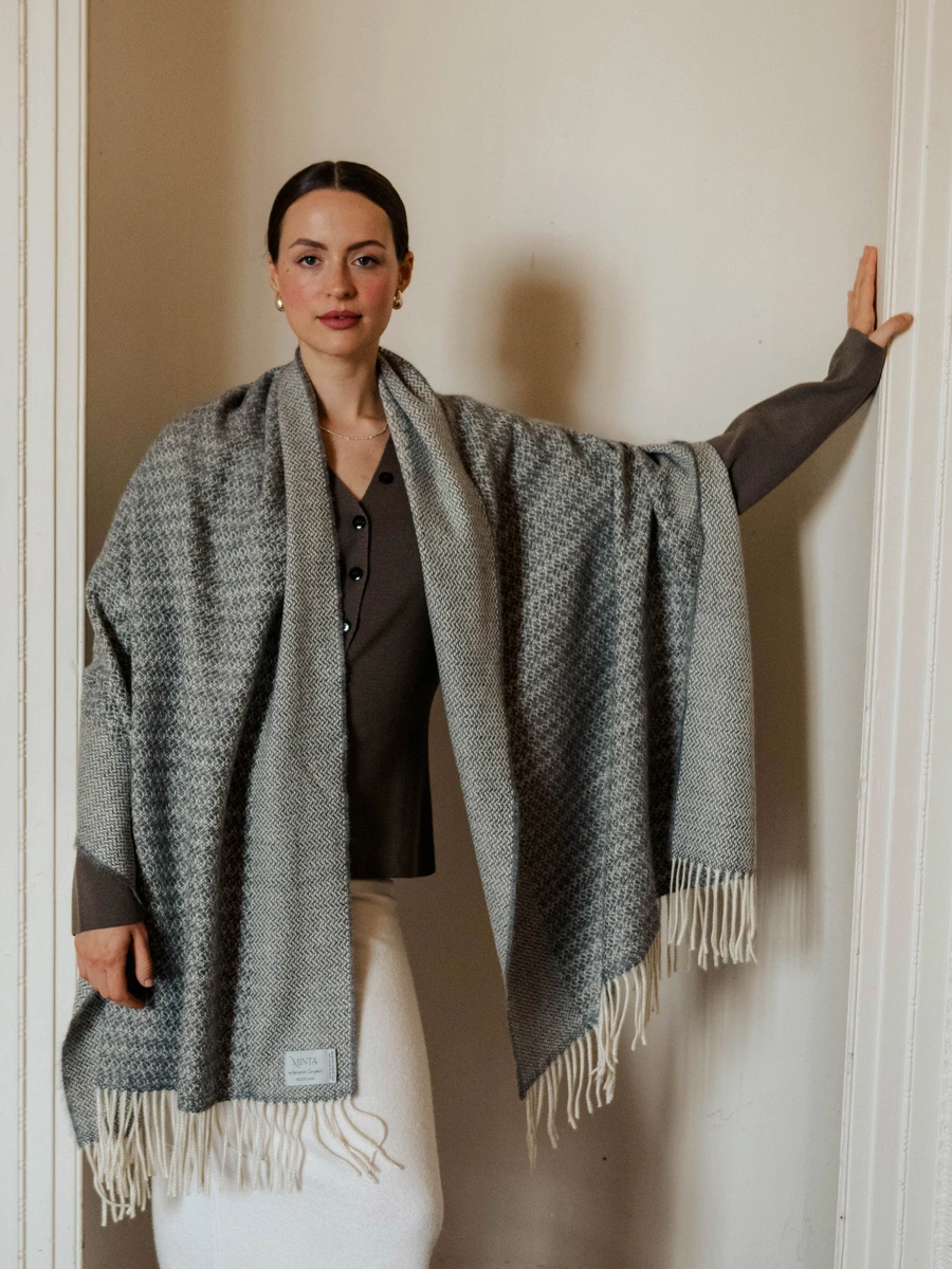 A woman standing against a beige wall, wearing a grey woven shawl with fringes, a dark grey top, and a white skirt.