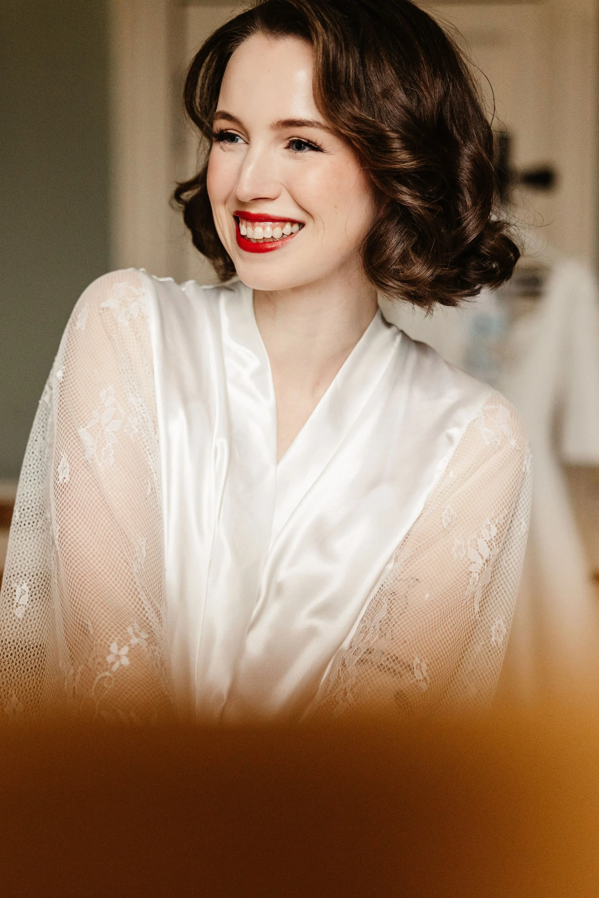 A young woman with short, wavy brown hair, smiling, wearing a silky white robe with lace sleeves.