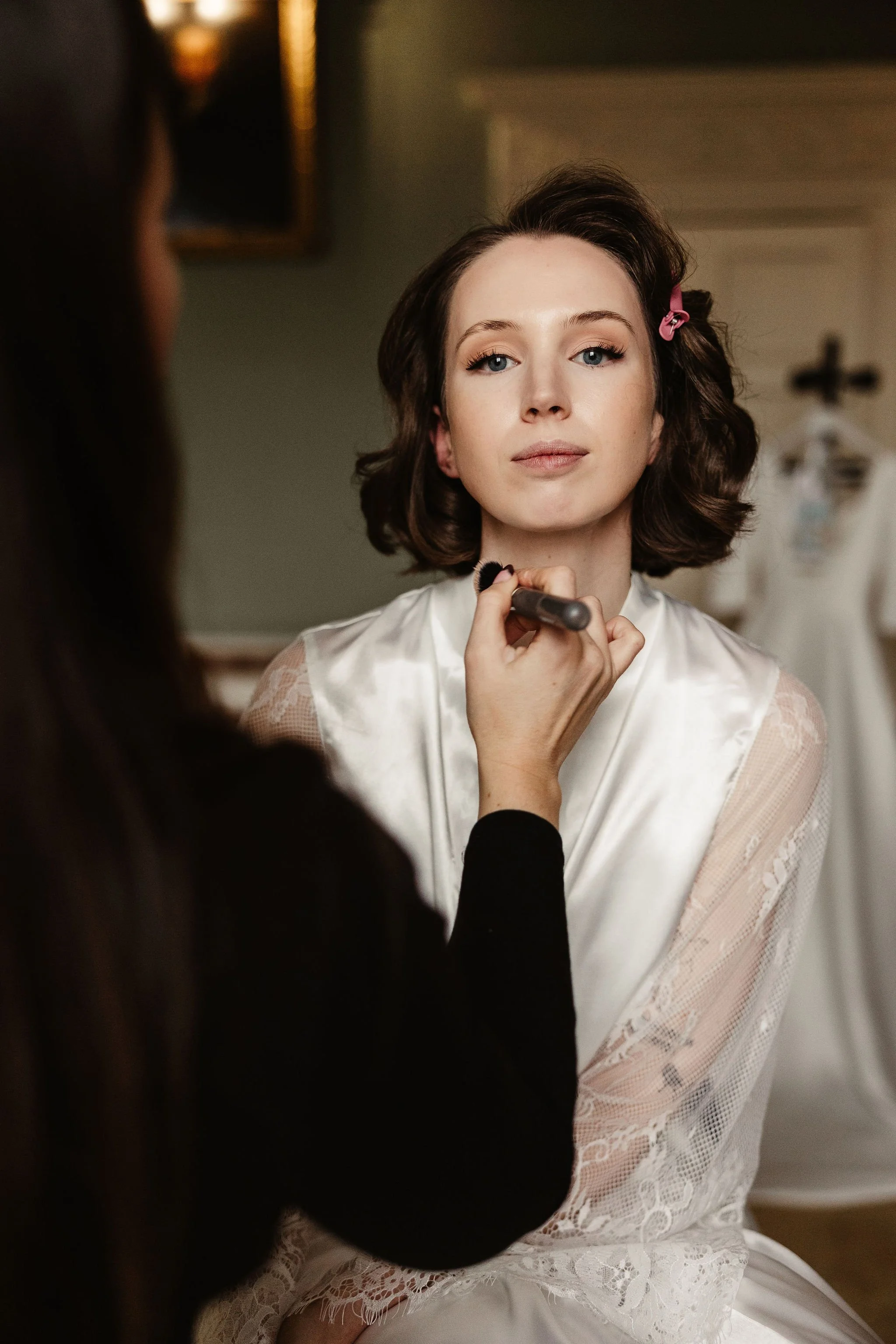 A woman is getting her makeup done by a makeup artist in a room with a dress hanging in the background. The woman has short, wavy brown hair and is wearing a white satin robe with lace on the sleeves.
