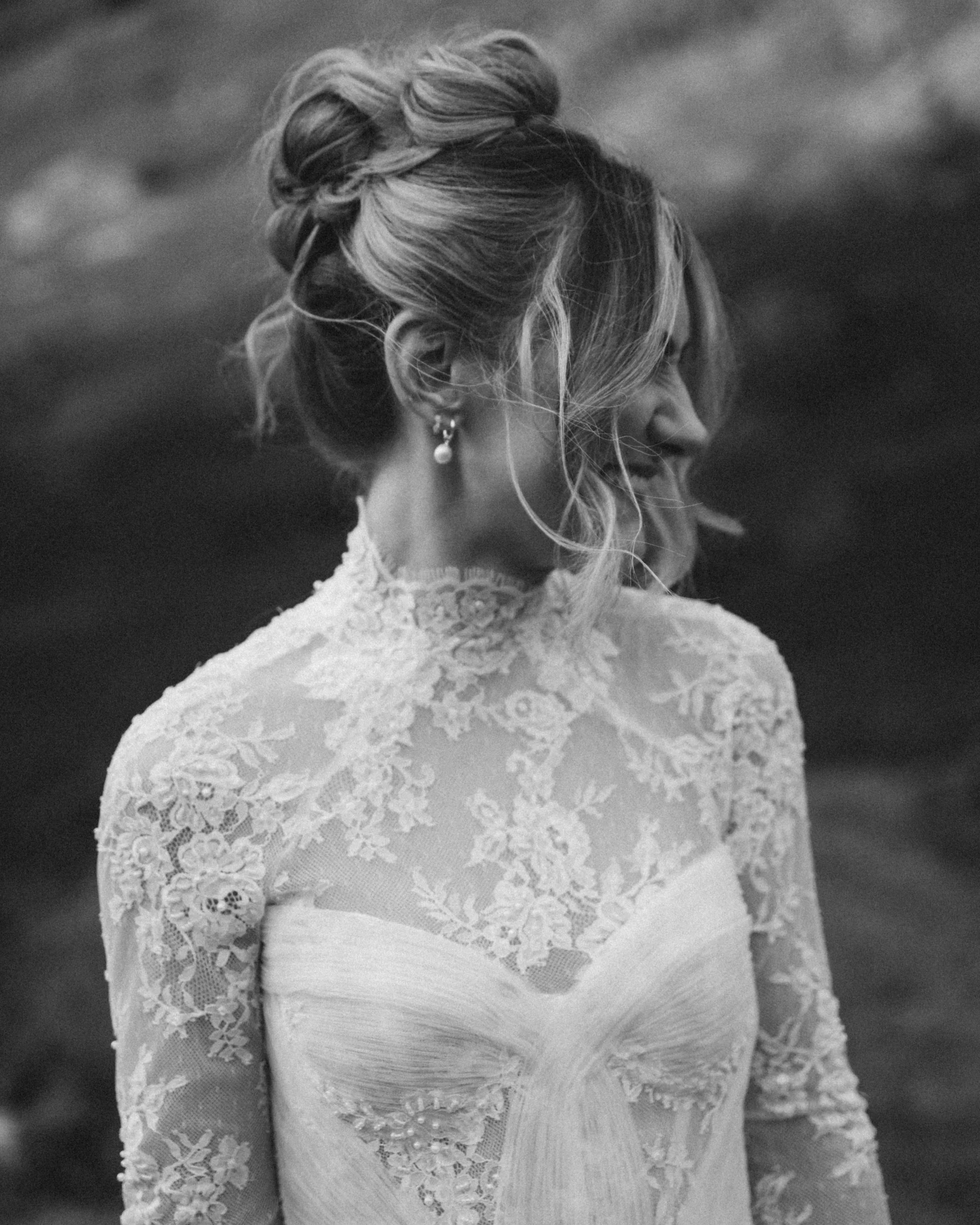 A woman in a lace dress with her hair styled in an updo, wearing pearl earrings, smiling and looking to the side.
