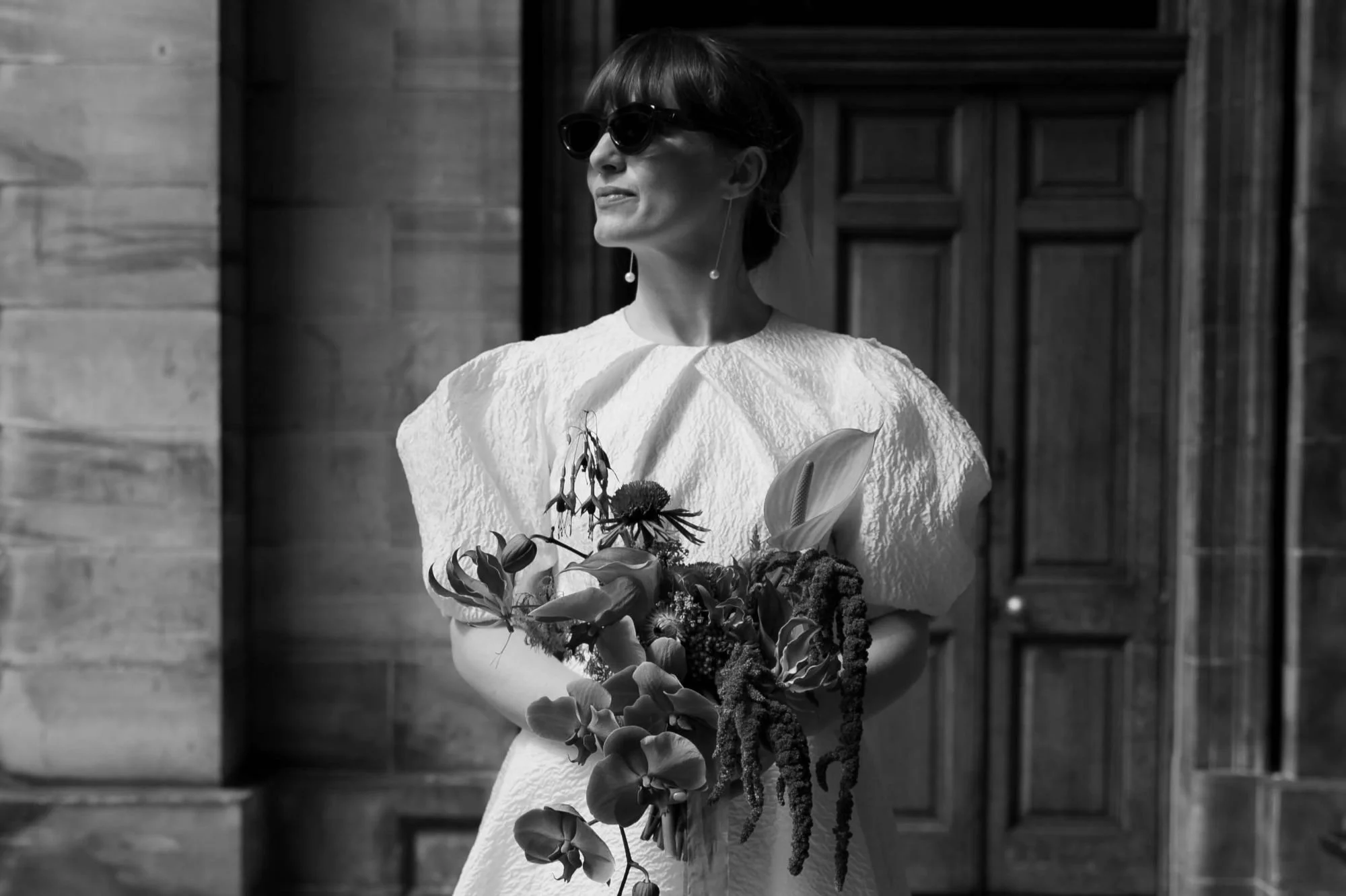 Black and white photo of a woman with short hair wearing sunglasses and earrings, holding a bouquet of flowers, standing in front of a wooden door.
