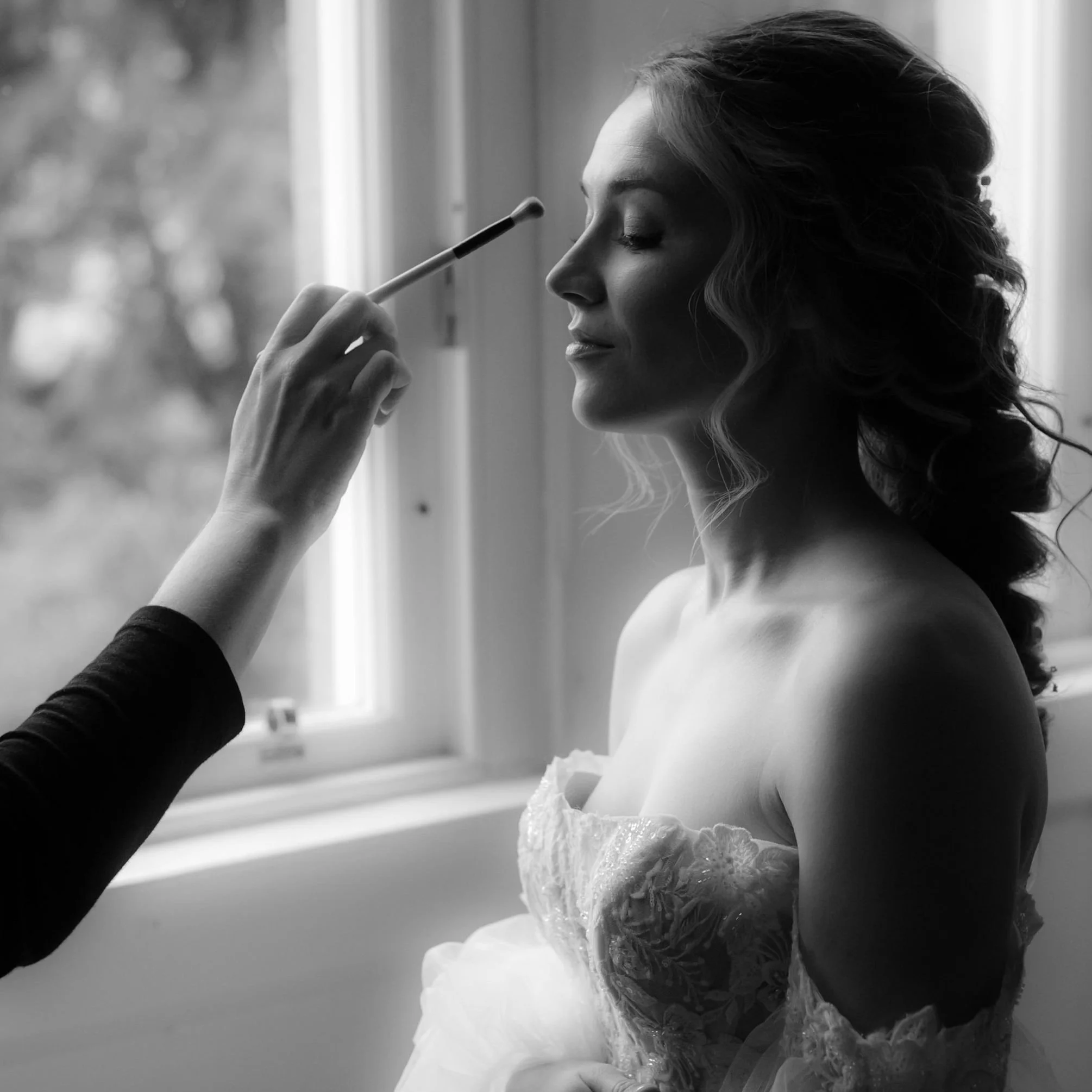 A woman with styled curly hair is having her makeup done by a makeup artist, with her eyes closed, near a window.