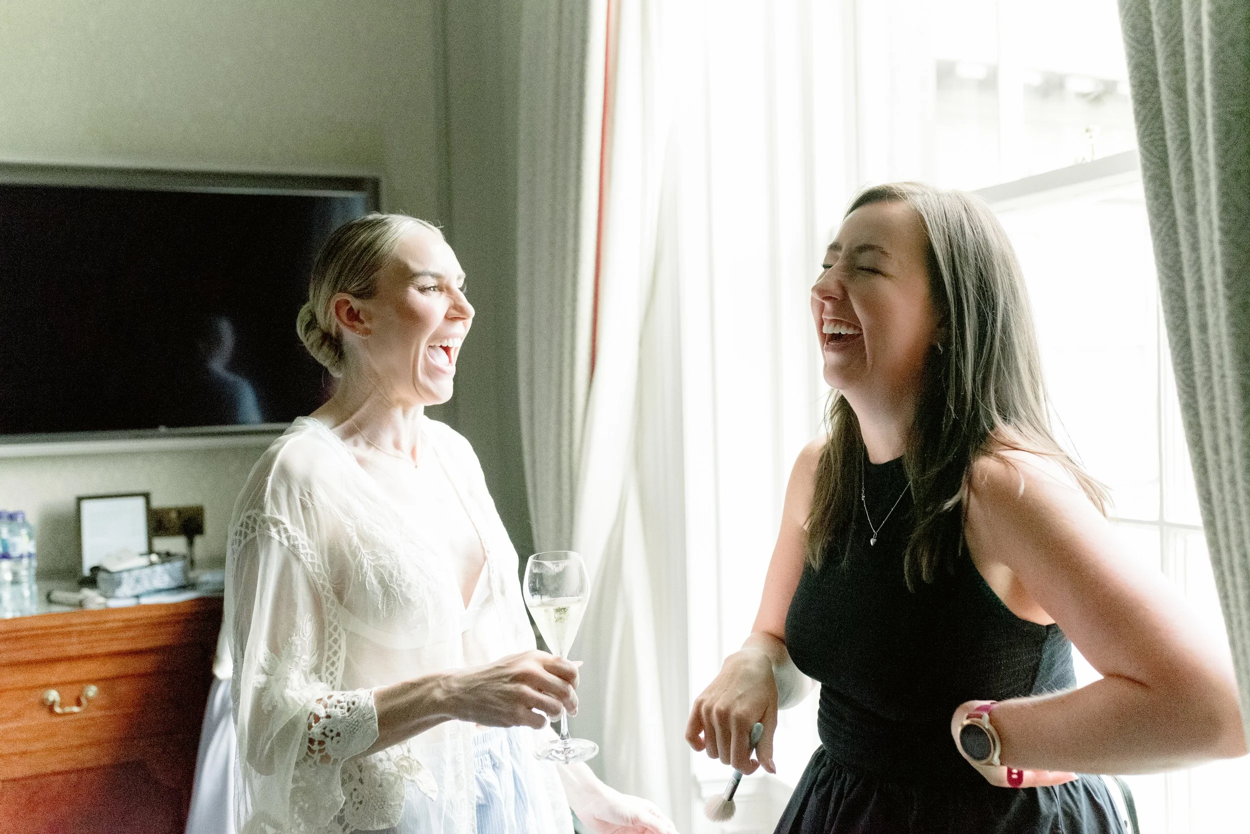 Two women laughing and sharing a joyful moment indoors. One woman is holding a glass of champagne, while the other is wearing a watch and a black dress.