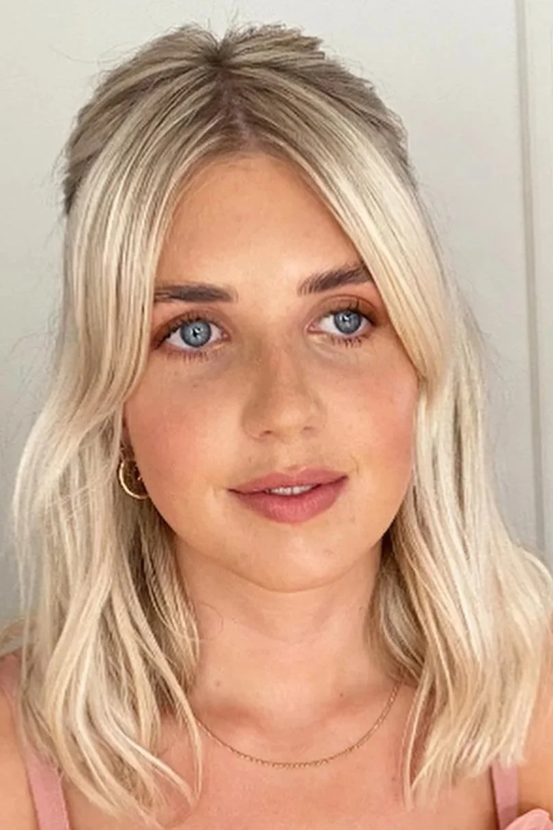 A young woman with blonde hair styled in loose waves and blue eyes, wearing gold hoop earrings and a delicate gold necklace, standing against a plain background.