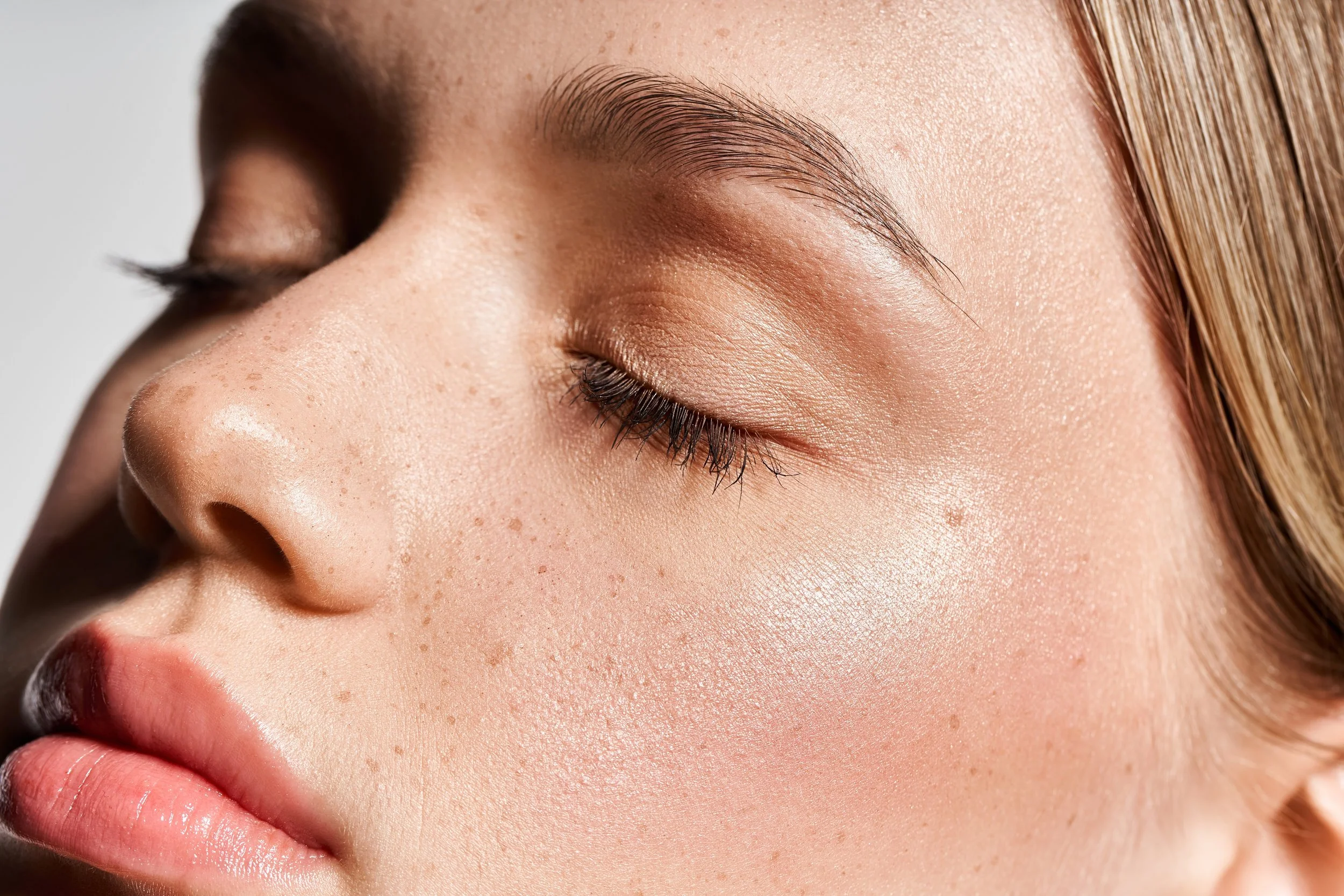 Close-up of a woman's face with closed eyes, showing smooth skin, natural makeup, and light freckles.