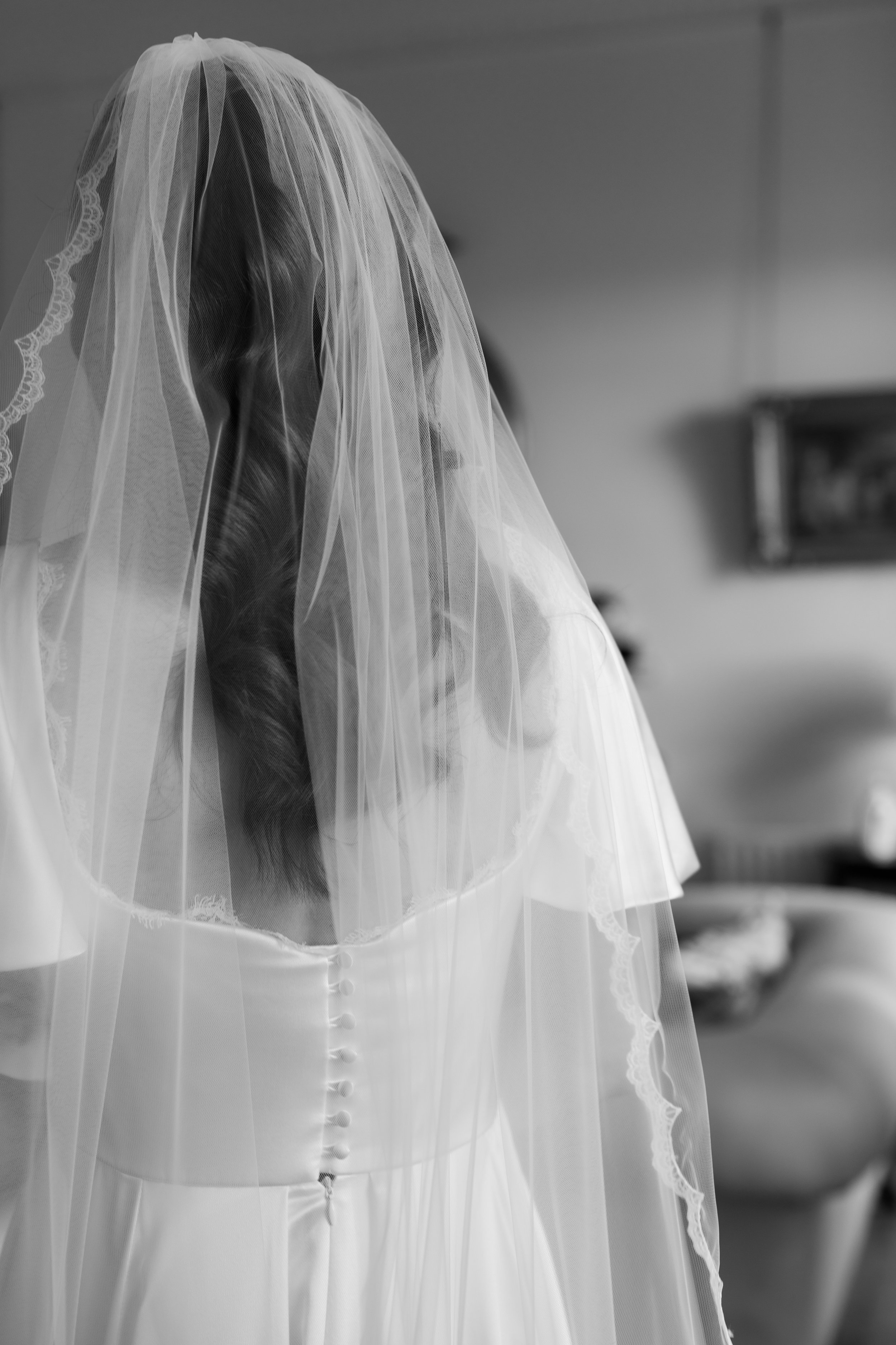 Black and white photo of a woman in a wedding dress with buttons on the back, wearing a veil, standing indoors.