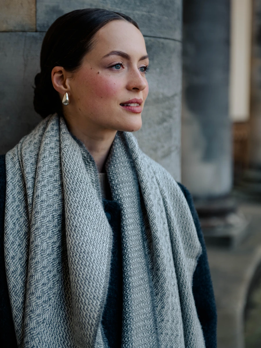 A woman with dark hair tied back, wearing earrings and a gray scarf, standing against a stone wall, looking to the side.