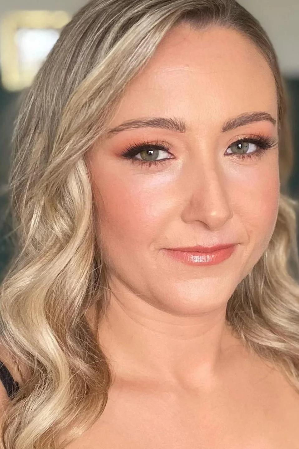 A woman with long brown hair in a white lace dress is touching her face in a room with wooden furniture and a mirror.