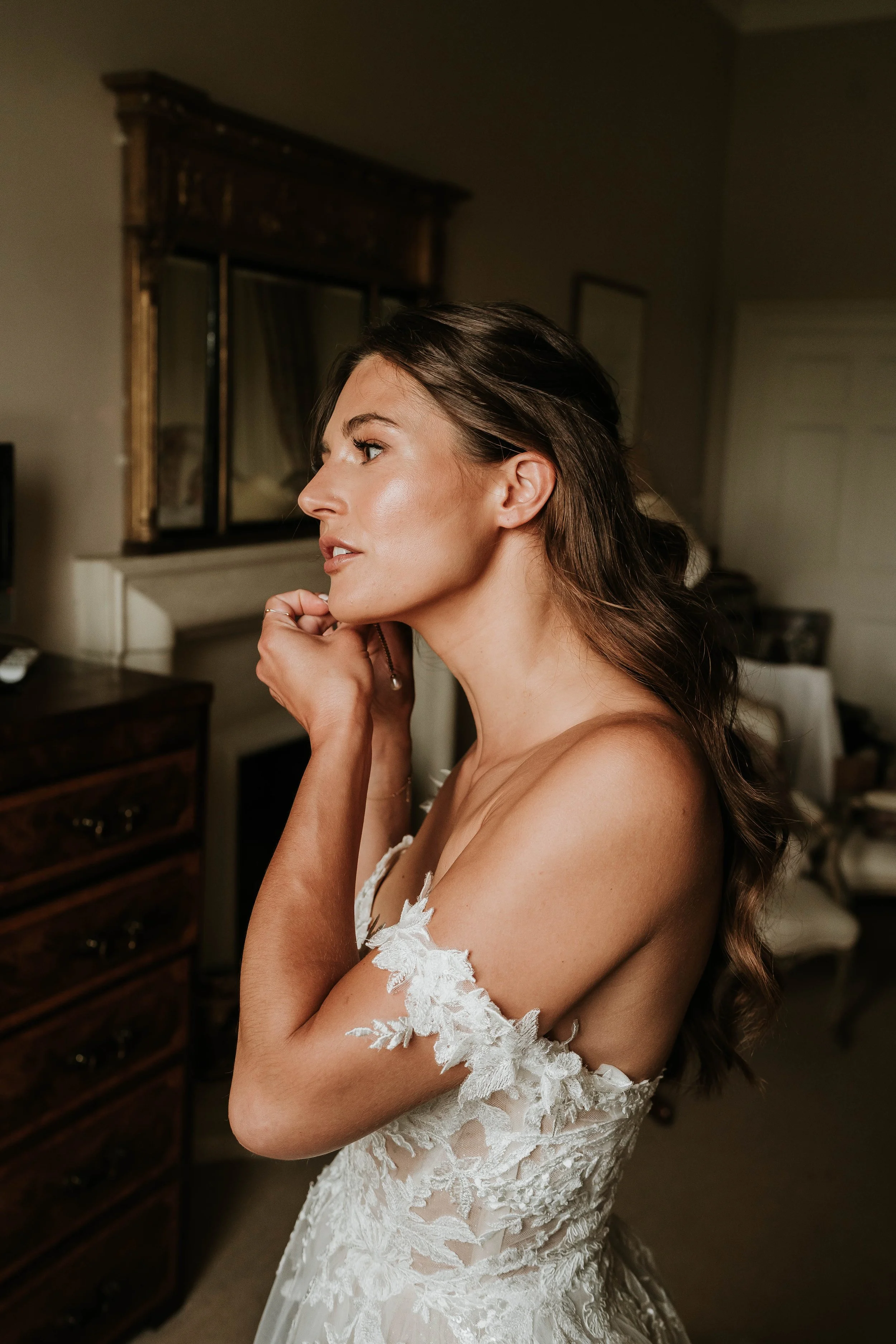 A woman with long brown hair in a white lace dress is touching her face in a room with wooden furniture and a mirror.