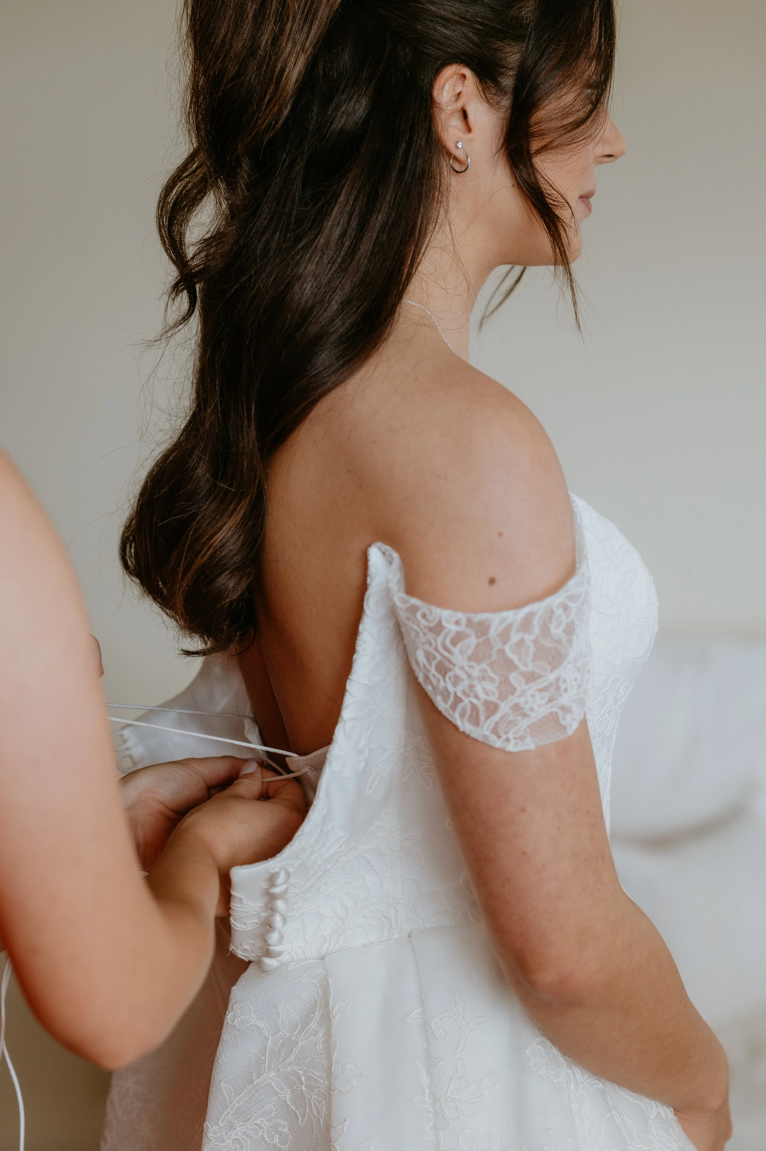 Woman in a white lace wedding dress with off-the-shoulder lace sleeves, having the back tied by another person.