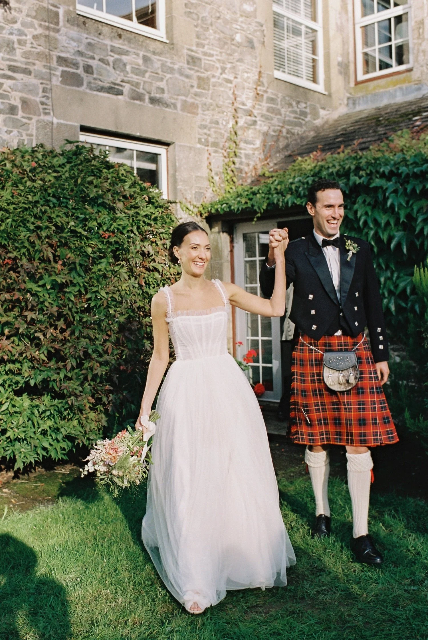 A bride and groom celebrating outdoors near a stone building, holding hands and smiling, with the bride in a white wedding dress and the groom in traditional Scottish attire.