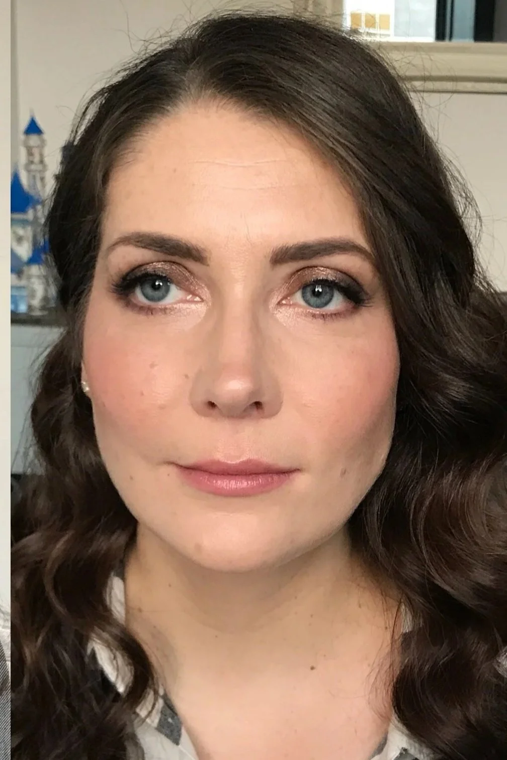 A woman with brown wavy hair and blue eyes looking at the camera, with a blurred background showing a blue and white castle model.