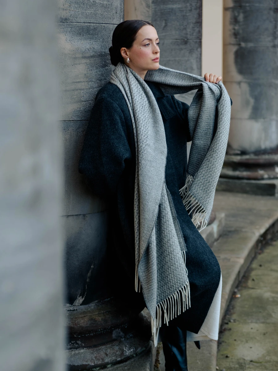A woman with dark hair slicked back, sitting against a gray stone wall, wearing a dark coat and a long gray scarf with fringed ends, looking thoughtfully into the distance.