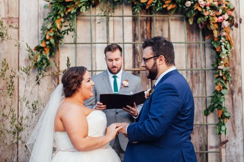 Bride & Groom Wedding Ceremony in front of a green & orange magnolia wedding arch. Eclectic Wedding at Big Daddy's Antiques Los Angeles, Groom in a blue suit, Della Curva Bride, Art & Soul Events