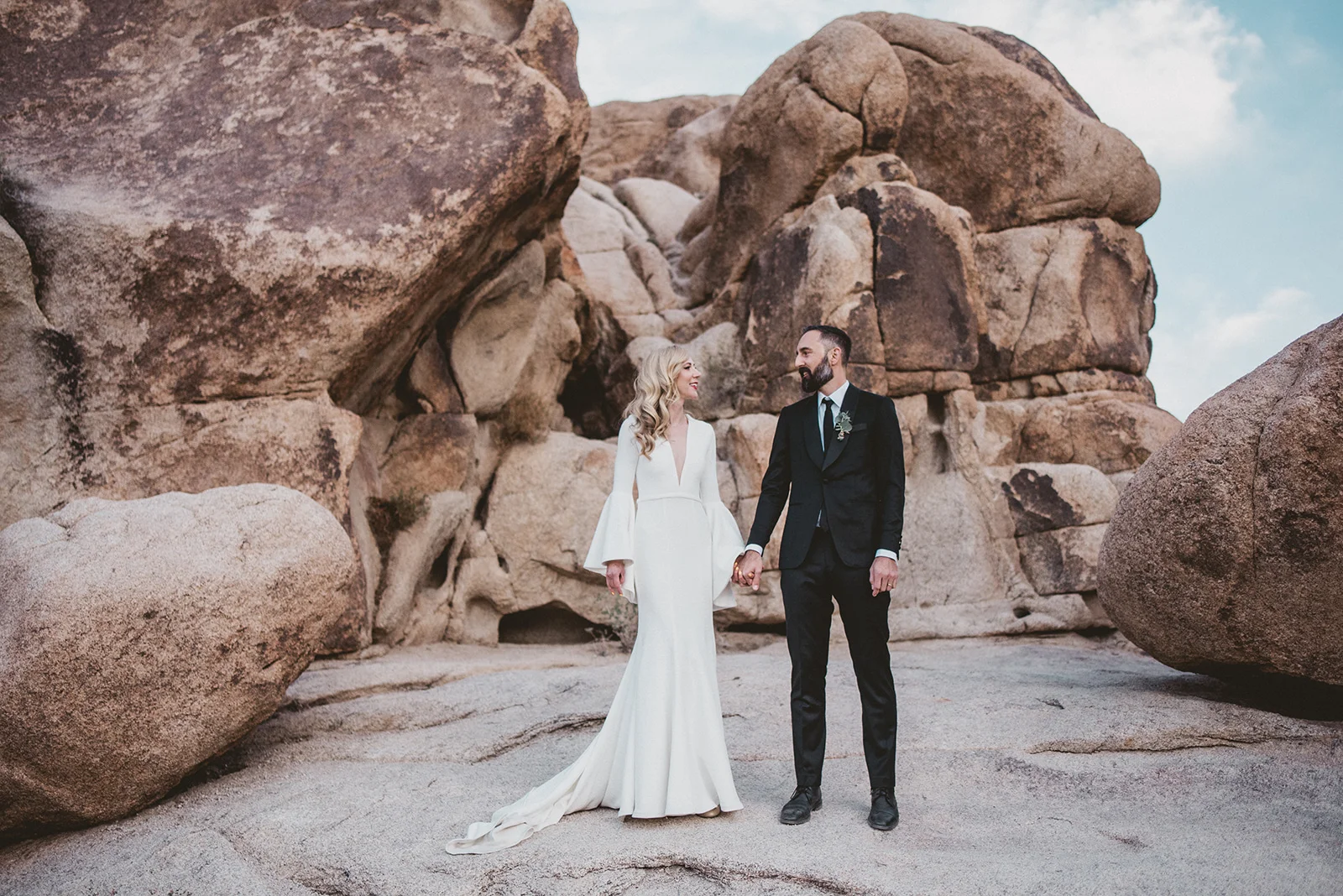 Epic Wedding portraits. Beautiful blonde bride in a Suzanne Harward The Bell Sleeve Wedding Dress at the Indian Cove Amphitheatre in Joshua Tree Ceremony, Hot bearded Groom in a black suit