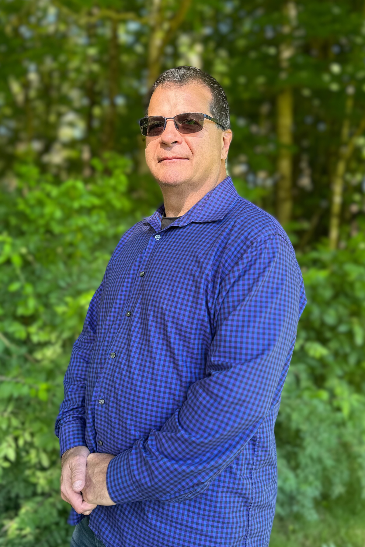 A smiling man wearing glasses, a red checkered shirt, and a black Columbia vest standing outdoors with trees and green foliage in the background.
