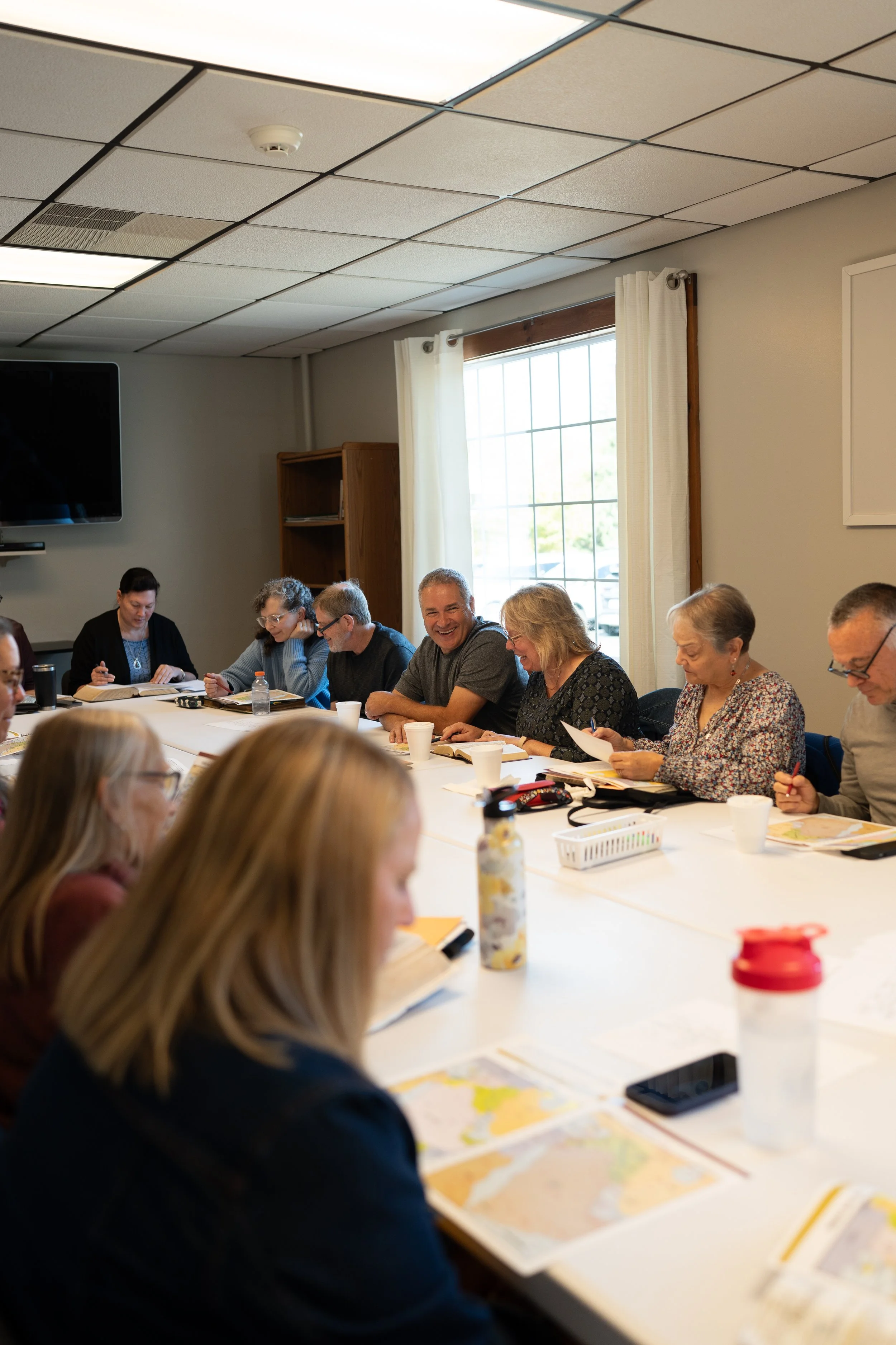 A group of fifteen people sitting around a large meeting table in a room with wood-paneled walls, smiling and facing the camera, with papers, drinks, and various personal items on the table.
