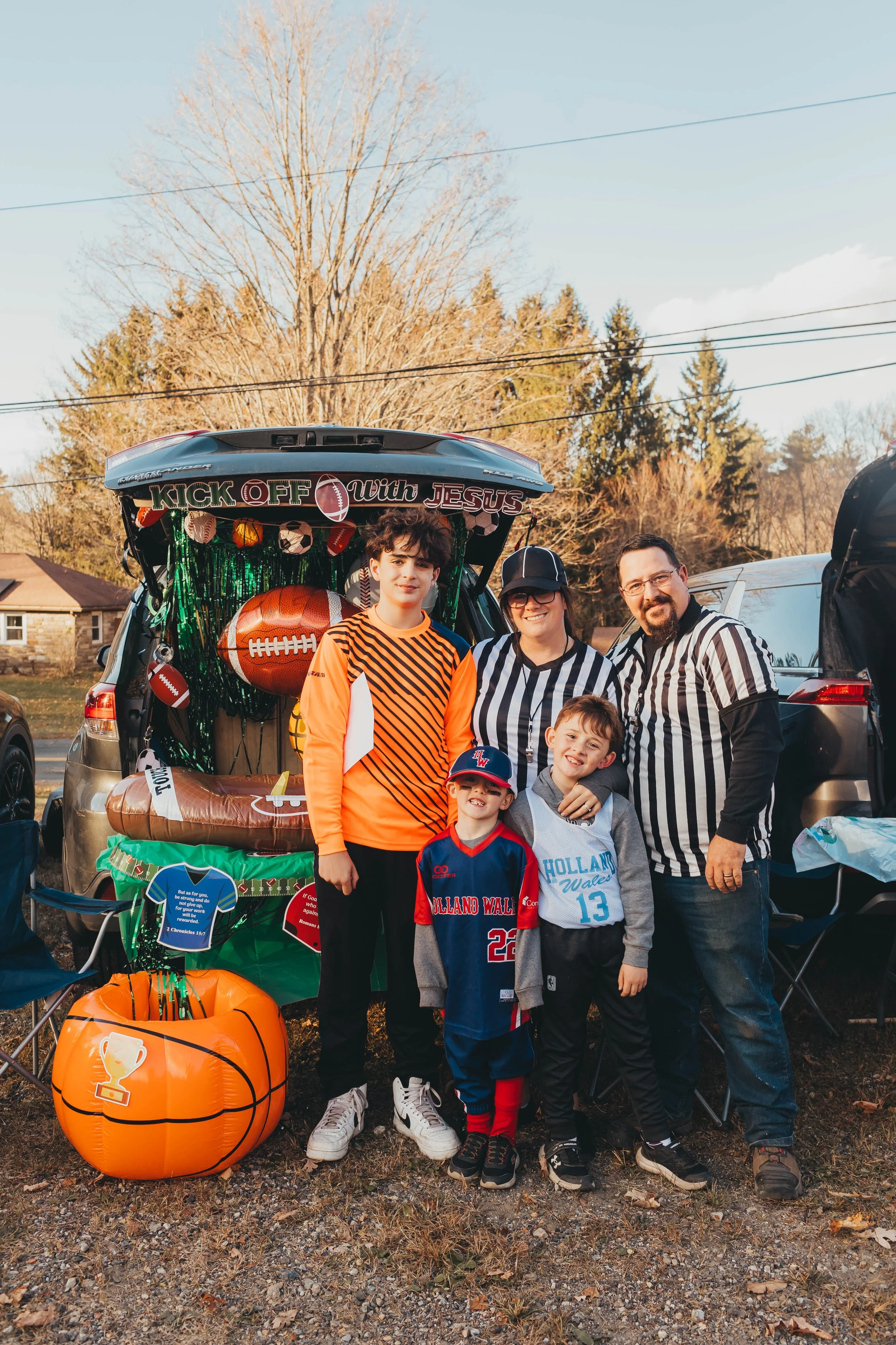 A woman dressed in a festive, striped shirt and pink skirt with a themed headband is handing out candy to children at a Halloween trunk-or-treat event, with a decorated car trunk in the background.