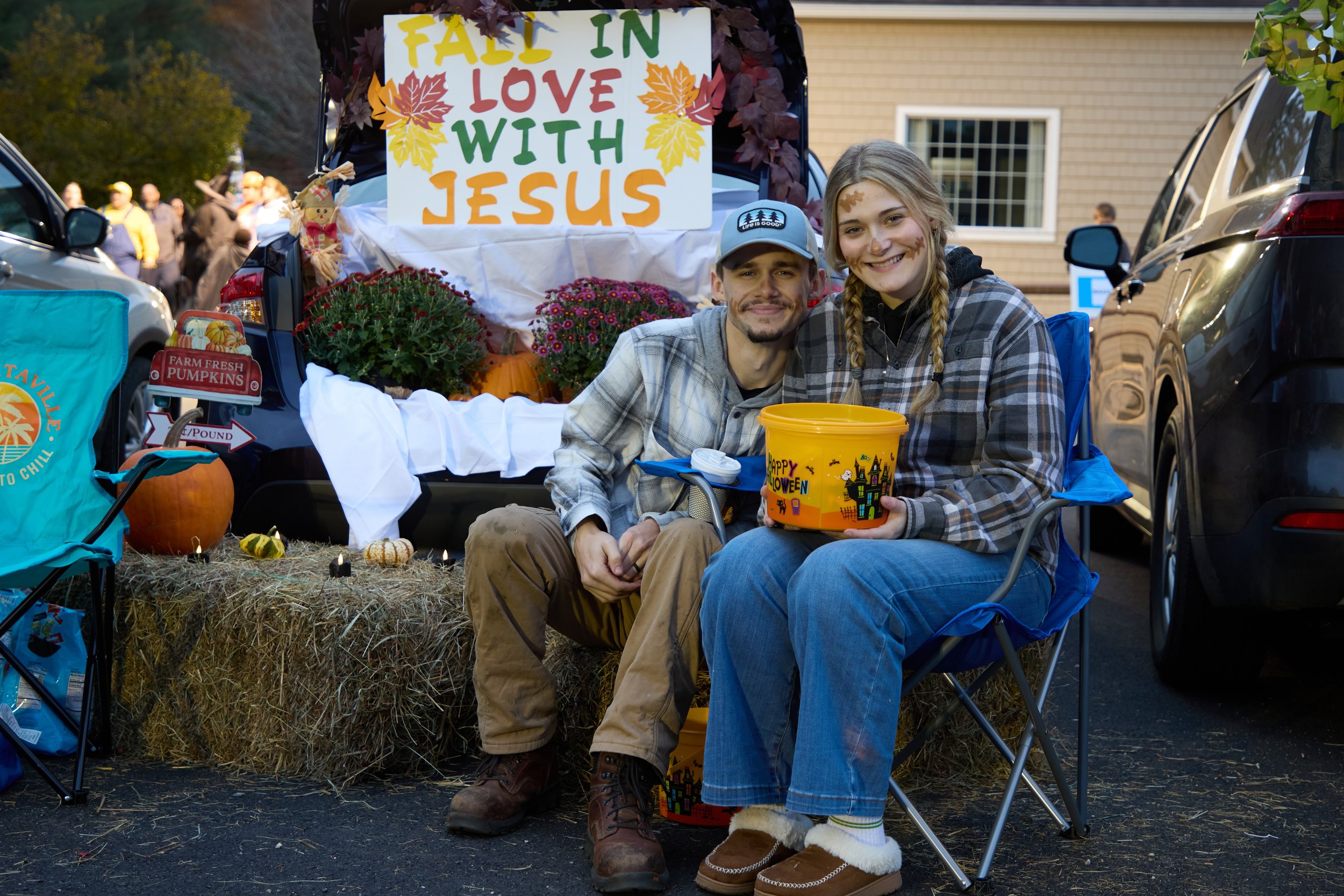 A woman dressed in a festive, striped shirt and pink skirt with a themed headband is handing out candy to children at a Halloween trunk-or-treat event, with a decorated car trunk in the background.
