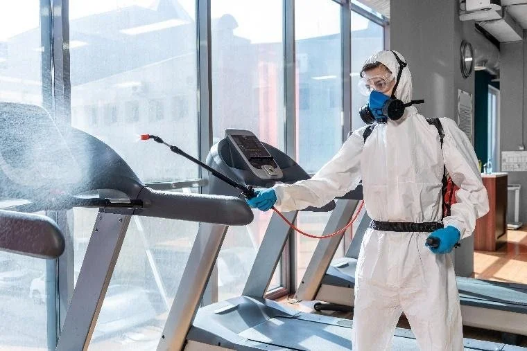 Person in full protective gear disinfects treadmill in a gym.