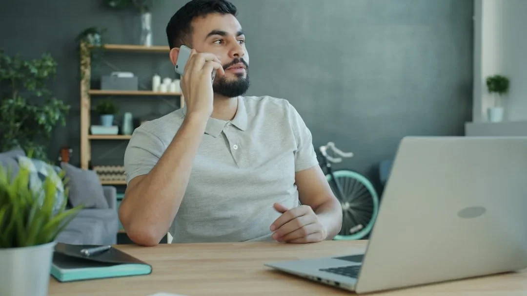 A man with dark hair and a beard, wearing a light gray polo shirt, sitting at a wooden desk in a modern room, talking on a smartphone. There is a laptop, a notebook, and a pen on the desk, with a green potted plant in the foreground and a bicycle in the background.
