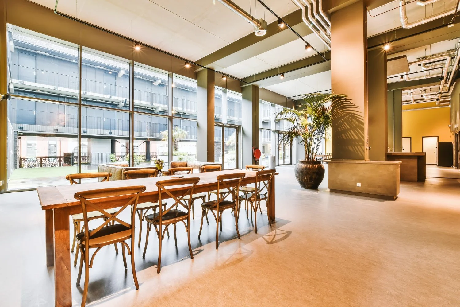 Modern interior space with large floor-to-ceiling windows, a wooden dining table with eight chairs, potted plants, and sunlight streaming in, with an industrial ceiling design.