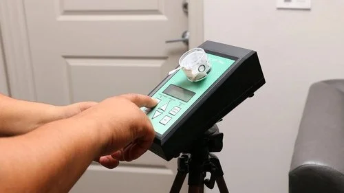 Person pressing buttons on a gray control device mounted on a tripod, with a small transparent container on top, in a room with a gray door and white walls.