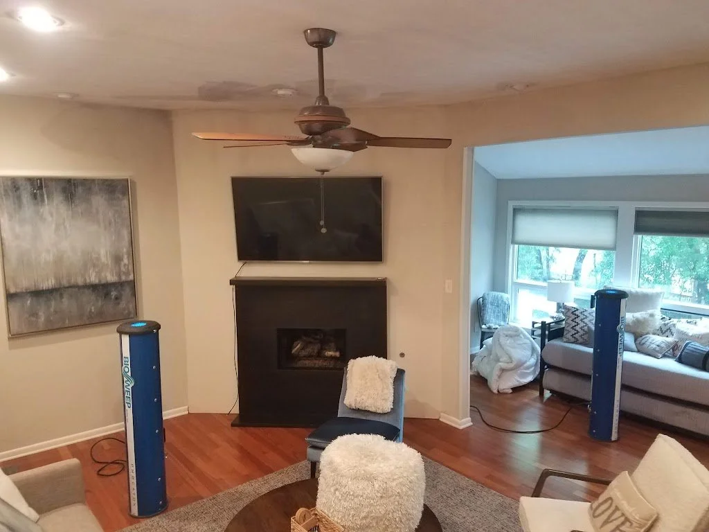 Living room with ceiling fan, wall-mounted TV, fireplace, beige walls, hardwood floors, and various furniture including a chair with a blanket, a couch with pillows, a small table with a fluffy cushion, and two large blue air purifiers.