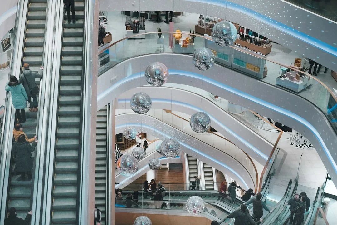 Multiple escalators in a multi-story shopping mall with shoppers riding them and decorative hanging spherical ornaments.