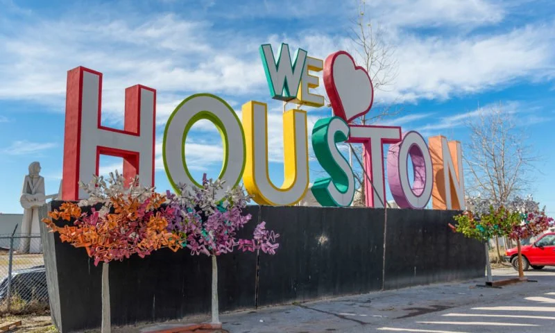 Colorful large sign reading "We ❤️ Houston" with flower arrangements at the base, against a blue sky with some clouds.