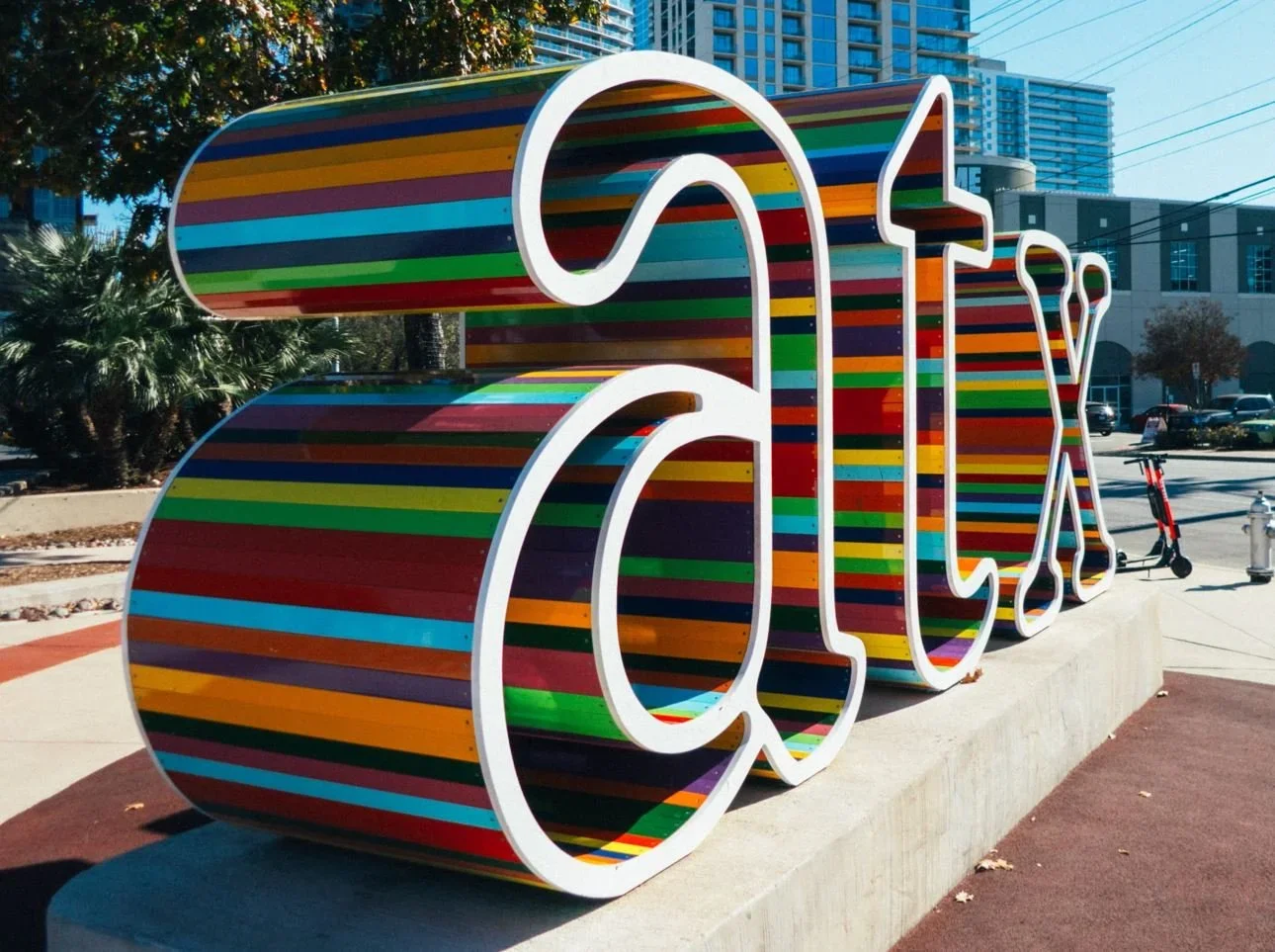 Colorful striped sculpture spelling out 'CITY' in outdoor urban setting with trees and buildings in background.