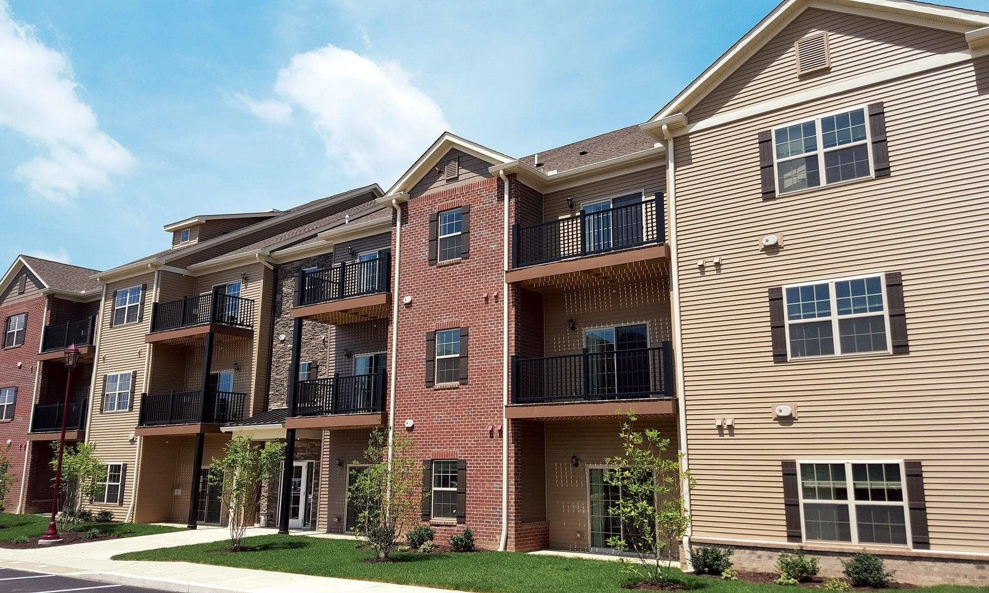 Multi-story residential apartment building with beige and brick exterior, black balconies, white trim, and a green lawn with young trees.