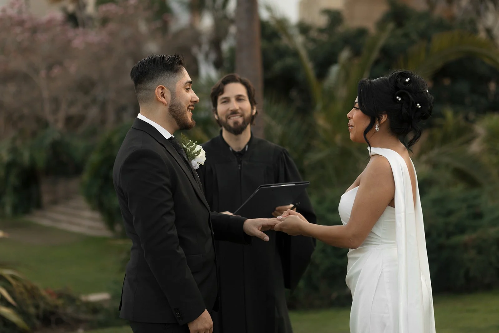 Couple holding hands with officiant present as groom places ring on bride during elopement ceremony at the Santa Barbara Courthouse Sunken Gardens