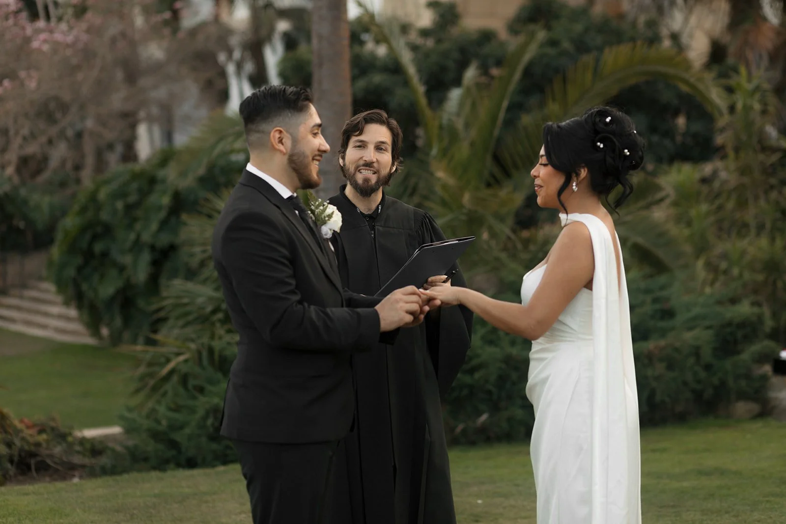 Couple holding hands with officiant present as bride places ring on groom during elopement ceremony at the Santa Barbara Courthouse Sunken Gardens