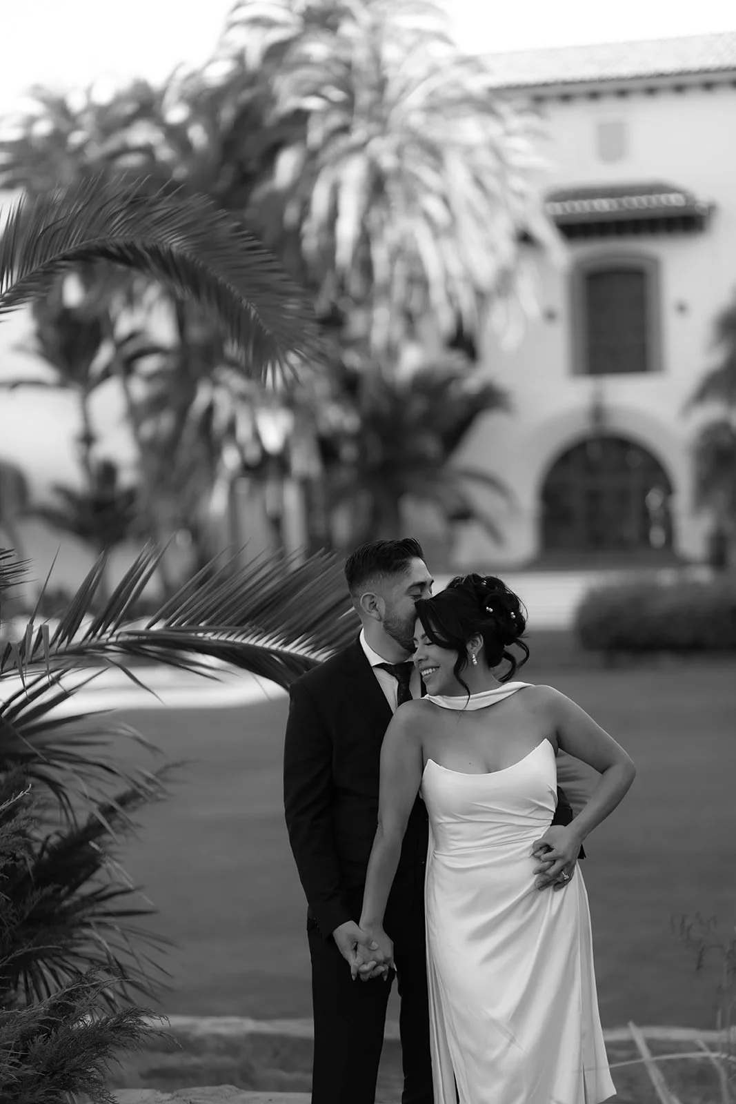 Black and white intimate elopement at Santa Barbara Courthouse gardens with groom embracing bride from behind and kissing her forehead