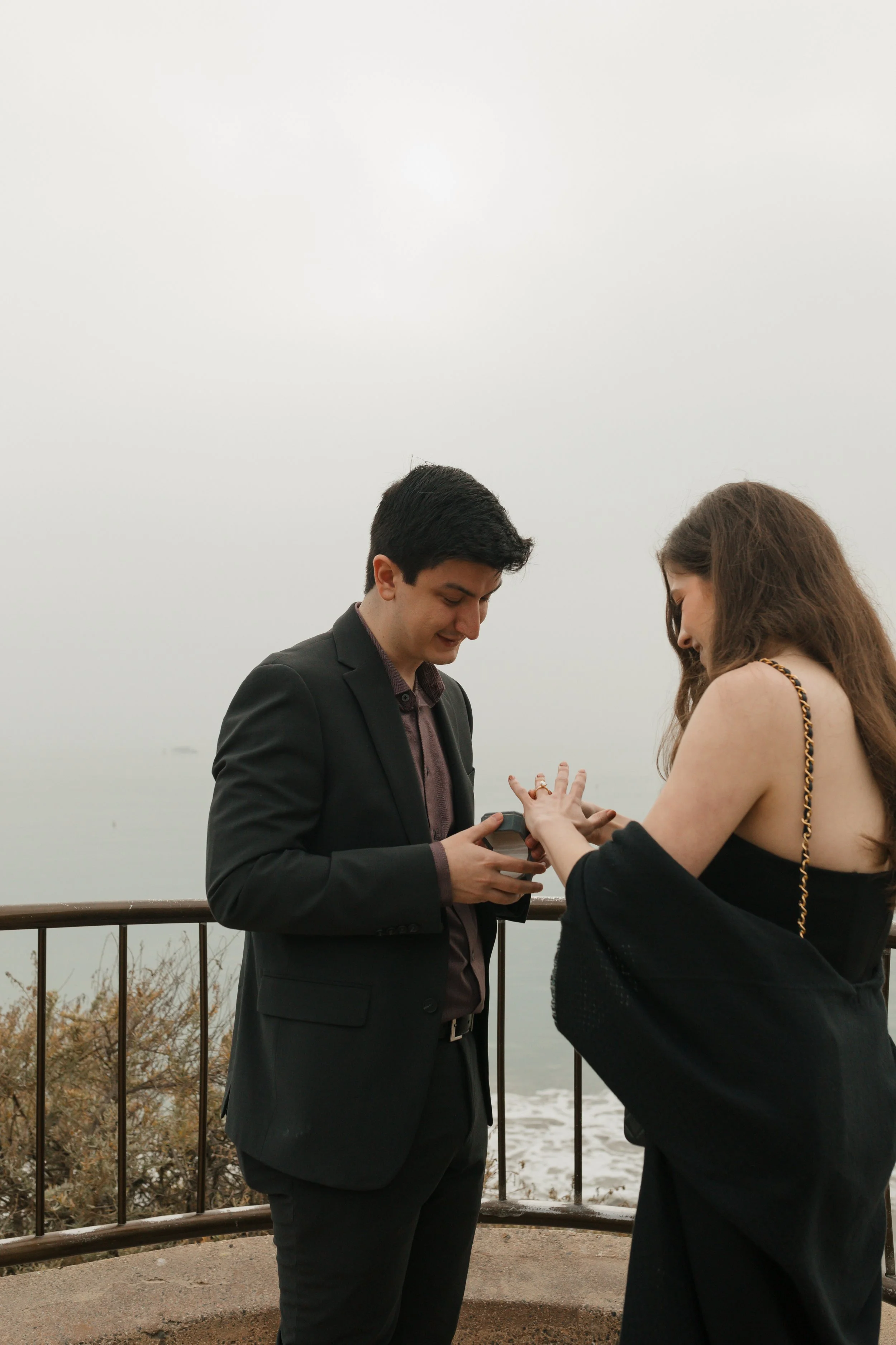 couple looking at ring as girl is putting on the ring from proposal at lookout in newport beach with the ocean in background