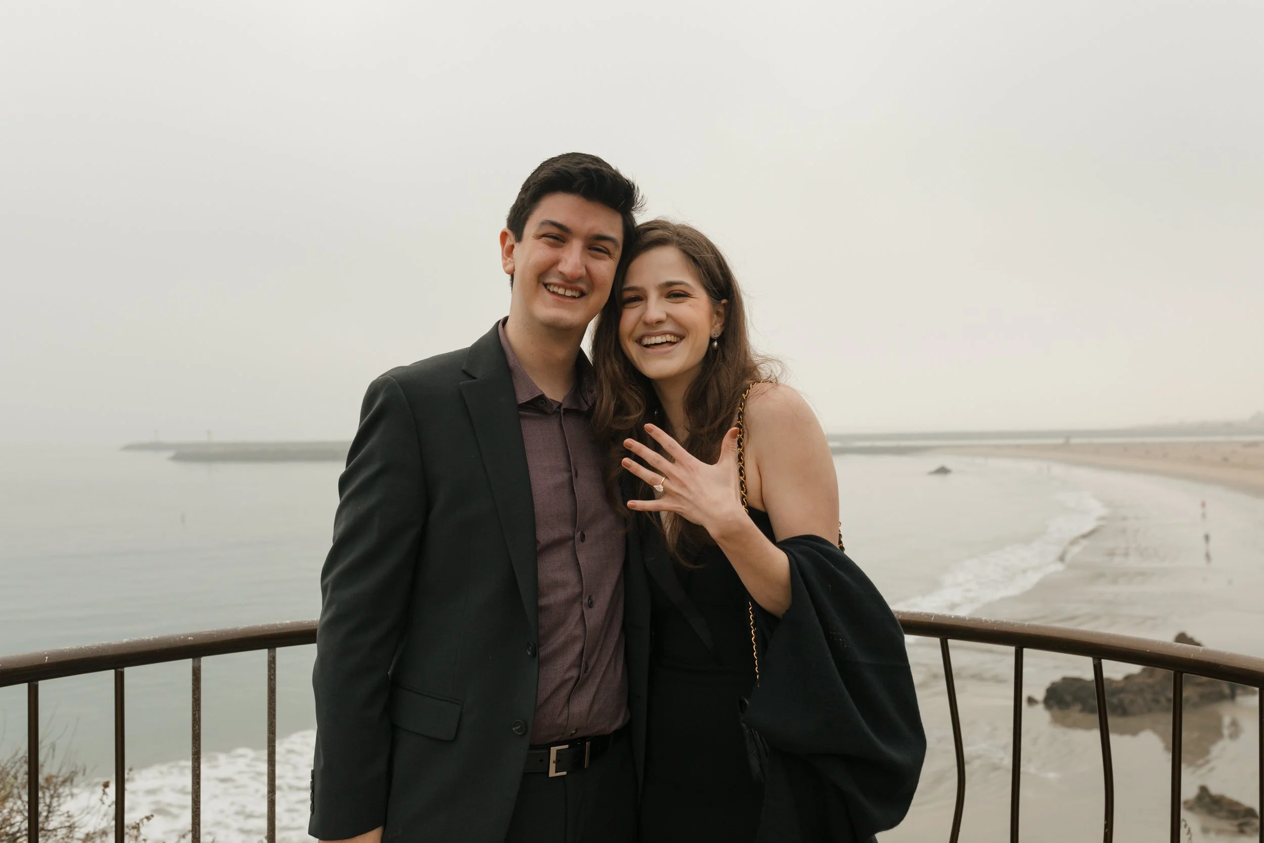 couple smiling at camera showing off the ring from the proposal at lookout in newport beach with the ocean in background