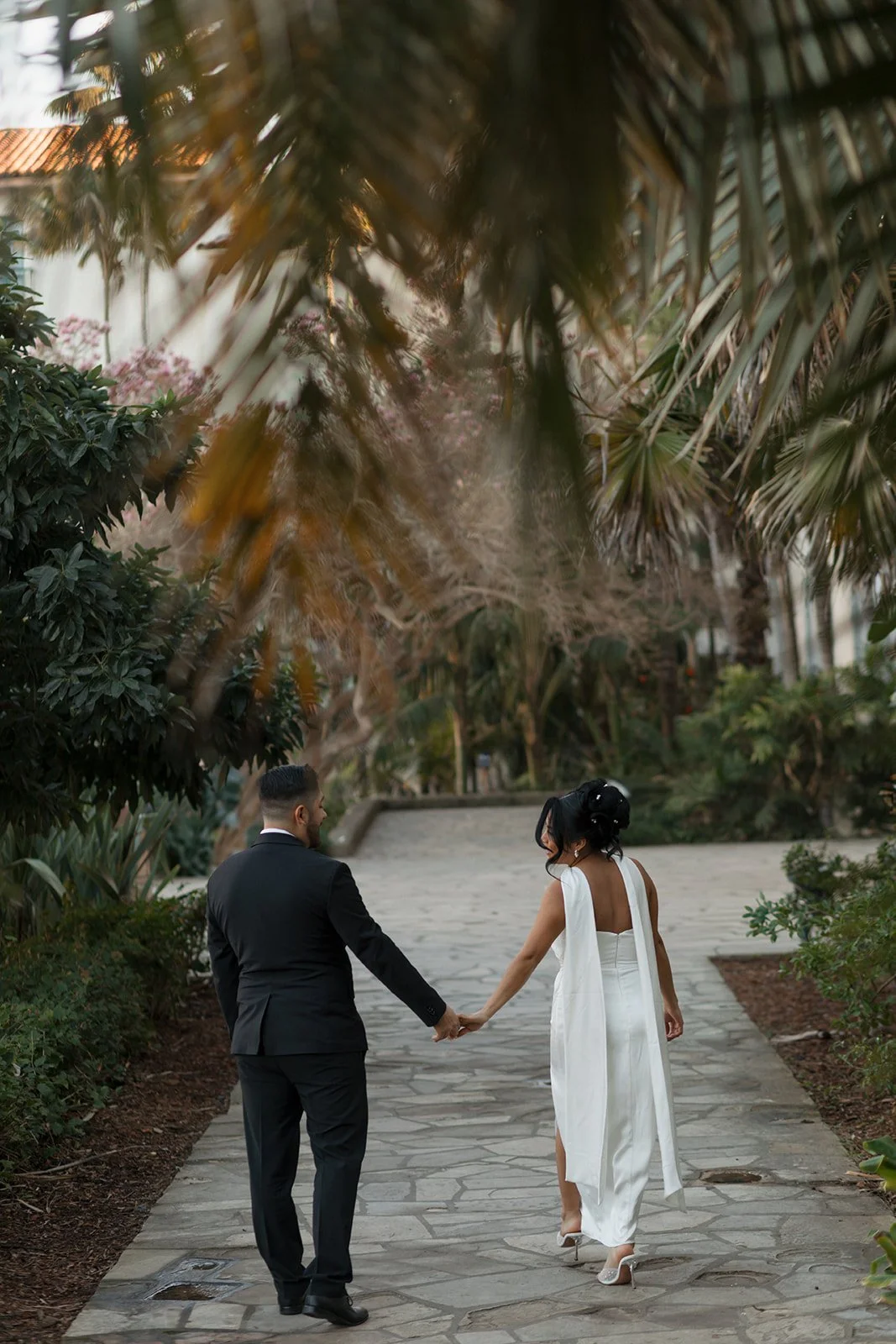 Artsy candid of couple holding hands while walking away at the Santa Barbara Courthouse Sunken Gardens, bride leaning in as they move together through the gardens