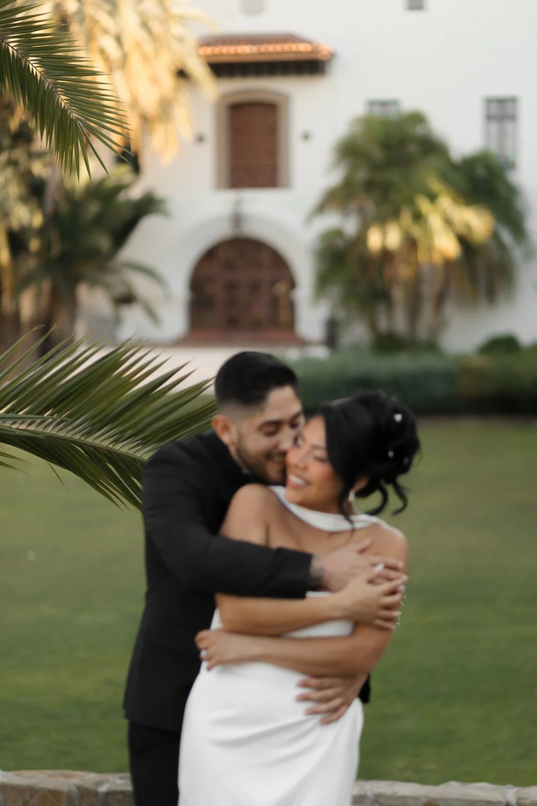 Groom embracing bride from behind in an intimate romantic moment at the Santa Barbara Courthouse gardens, both standing close together surrounded by lush greenery, capturing a soft and affectionate elopement scene.